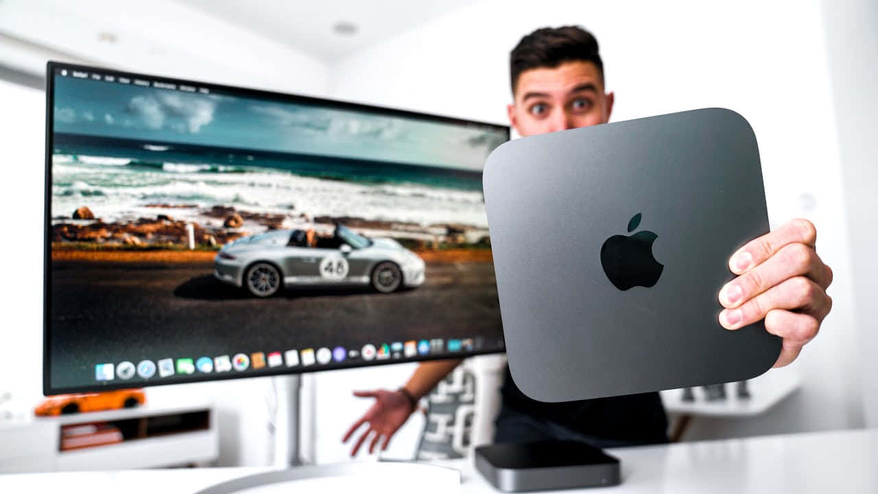 男子在顯示器前手持 Mac mini 電腦，顯示器顯示海灘上的賽車。