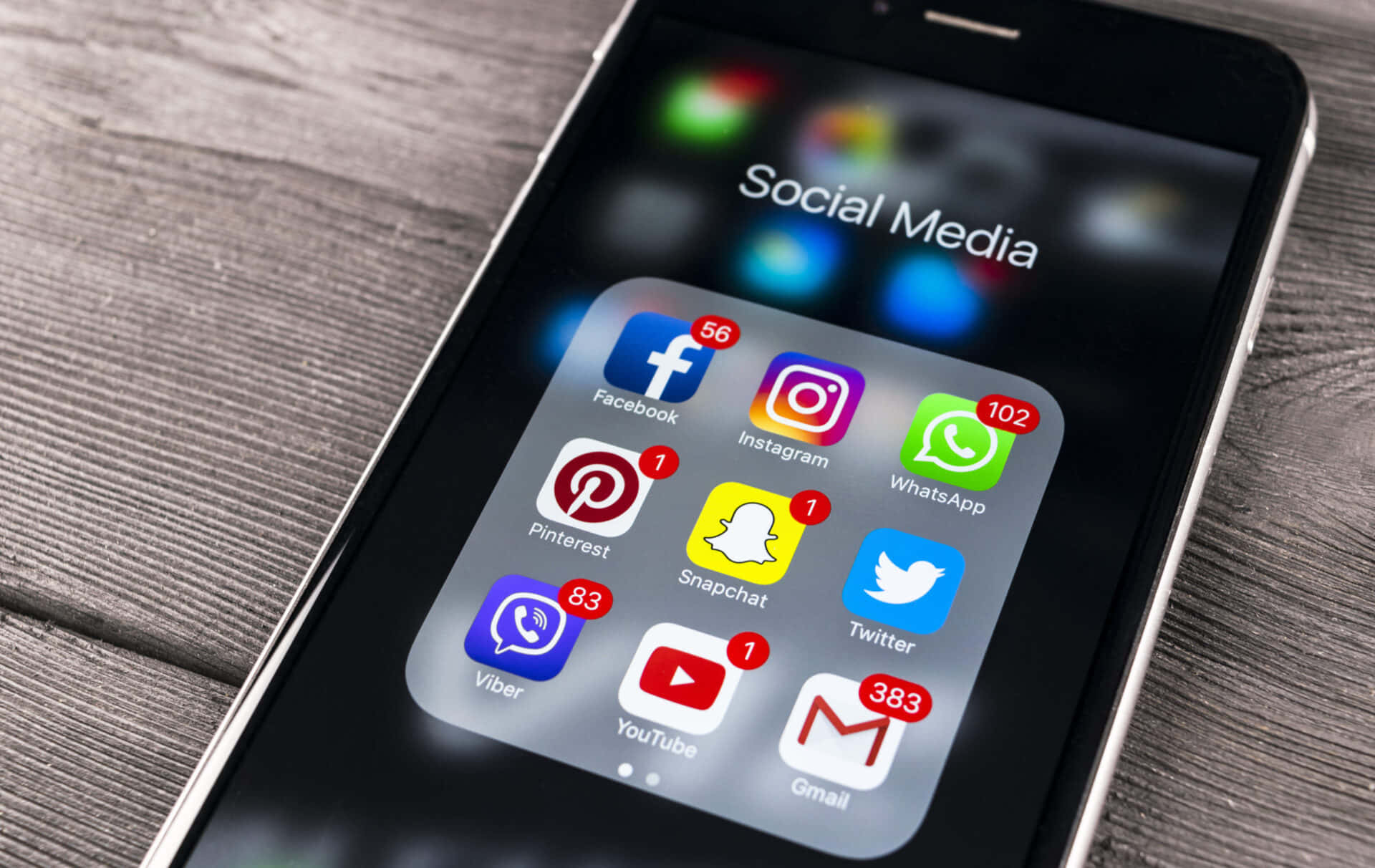 Social Media Logos 桌布 壁紙 — 木質表面上智能手機的特寫，顯示一個帶有通知徽章的應用程式圖標的「社交媒體」資料夾。