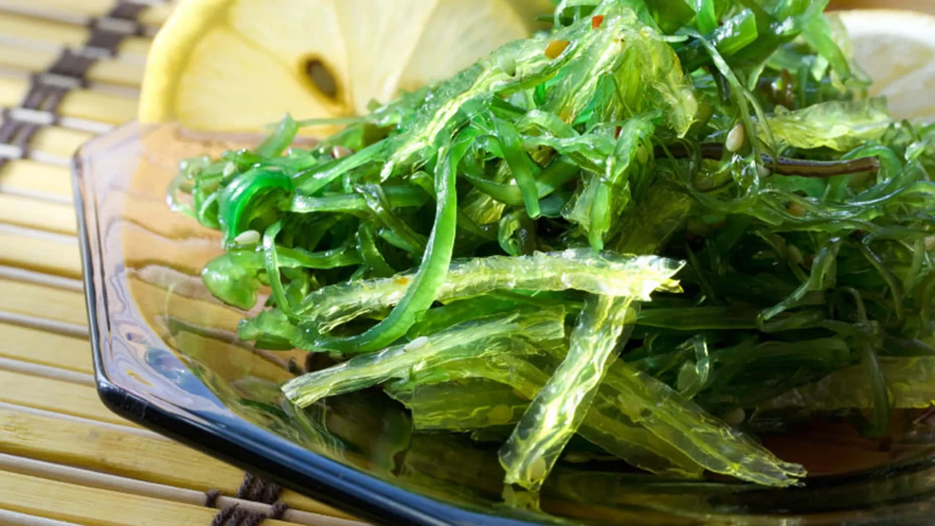 未受干擾的海灘風光，大自然的純淨之美 壁紙 — 一碗充滿活力的綠色海藻沙律在深色玻璃碗中閃閃發光，旁邊是竹蓆上的檸檬片。