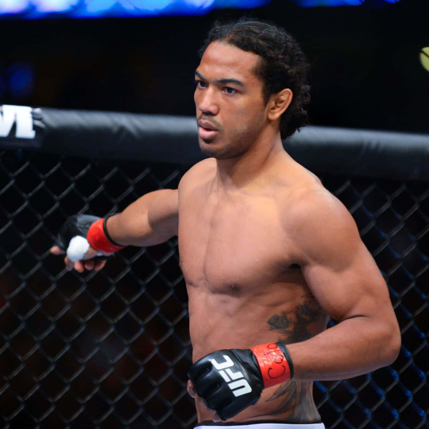 本·亨德森於UFC 150在百事中心展現實力 壁紙 — 一位肌肉發達的混合武術家戴著紅黑手套，在明亮的競技場燈光下警覺地站在UFC八角籠內。