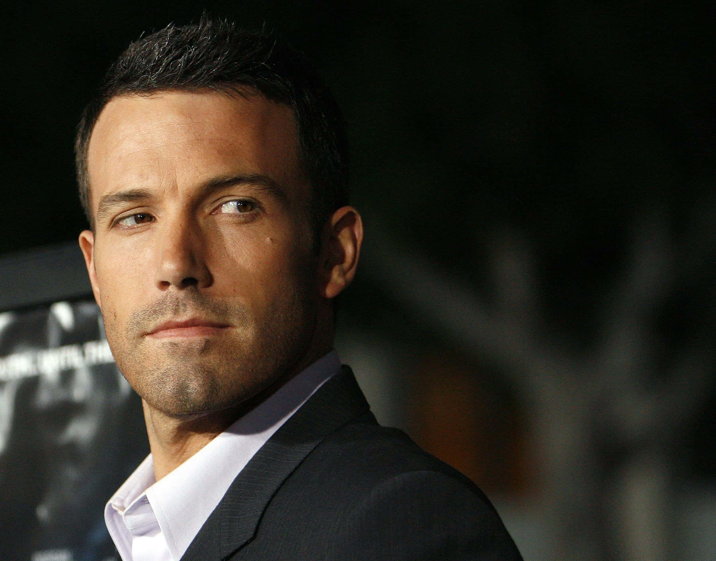 本·阿弗萊克於拍攝新作時稍作休息 壁紙 — 演員賓·艾佛力（Ben Affleck）的頭像特寫，身穿深色西裝和白色襯衫，背景模糊昏暗。