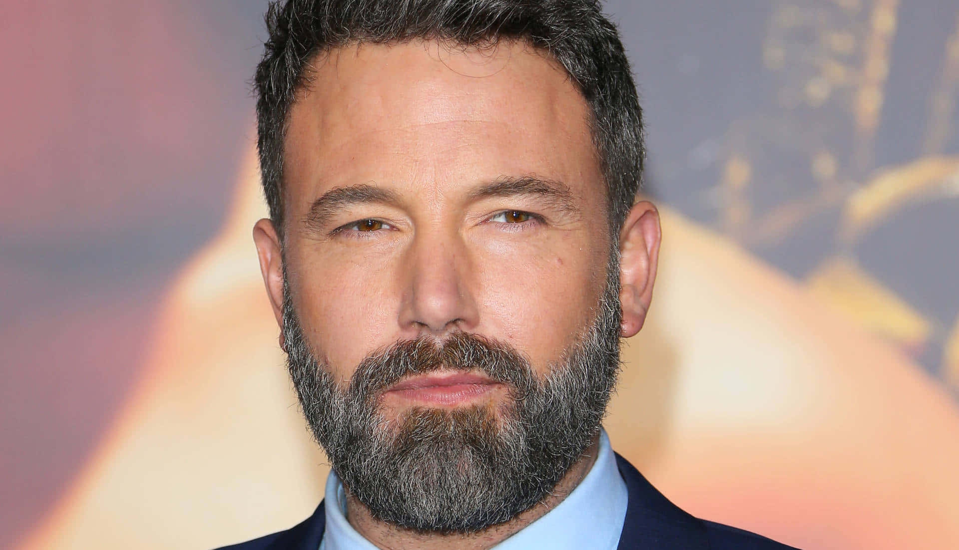 本·阿弗萊克望向城市天際線 壁紙 — 演員賓·艾佛力（Ben Affleck）的頭像特寫，留著滿臉鬍鬚，身穿西裝和淺藍色襯衫，背景模糊，色調溫暖。