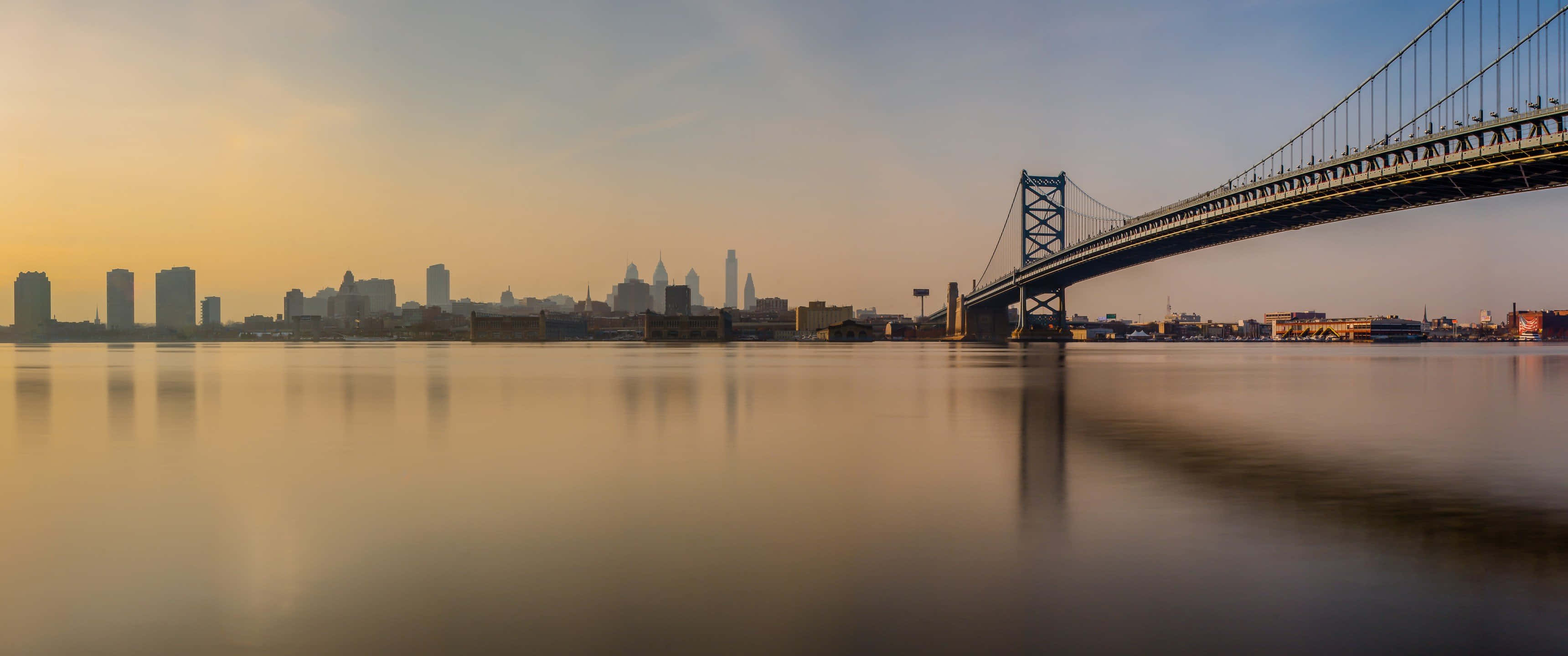 本傑明·富蘭克林大橋 3440x1440p 背景 壁紙 — 長時間曝光捕捉到日落時的班傑明·富蘭克林大橋，其倒影在平靜的水面上閃爍，遠處是金色的城市天際線。