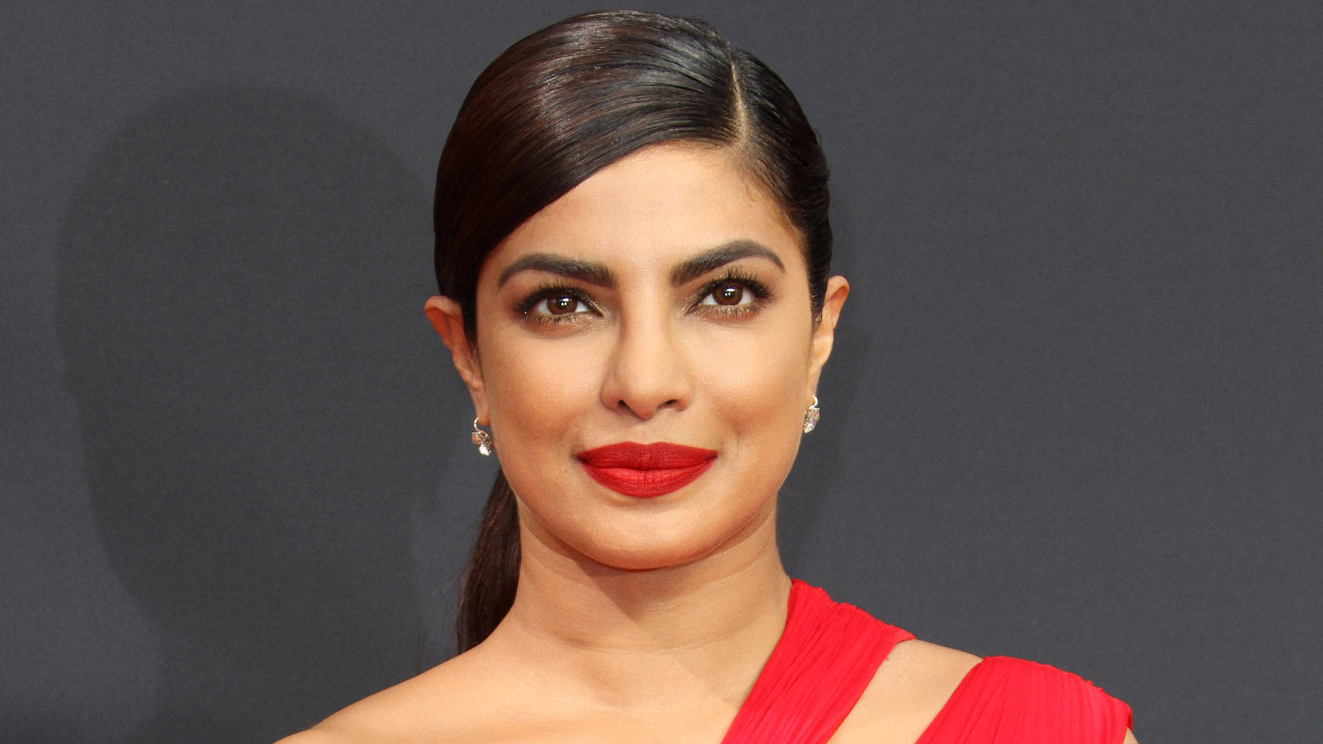 朴雅卡·喬普拉紅唇造型 壁紙 — Priyanka Chopra 喺深灰色背景前自信咁笑住，着住一條大膽嘅紅色連身裙。