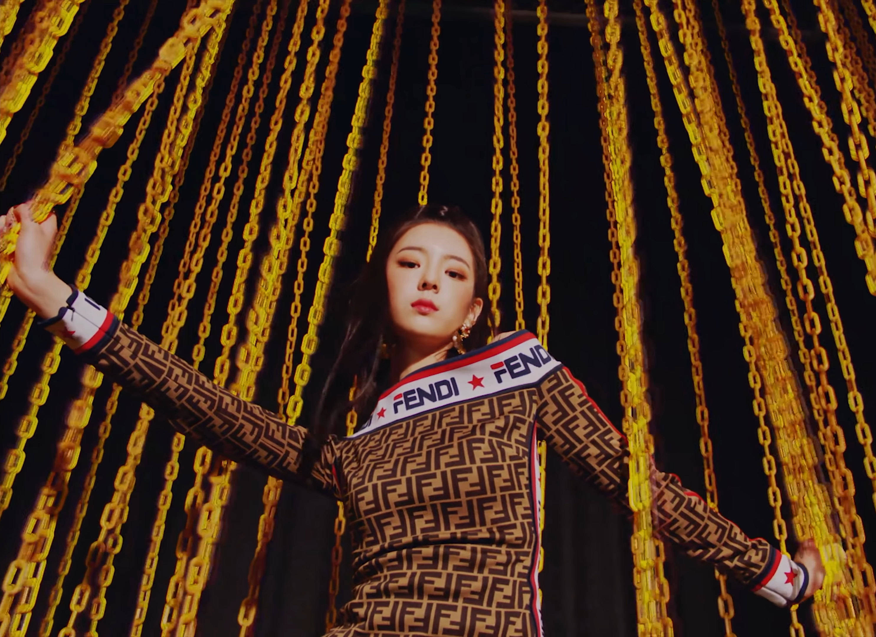 李婭低角度攝影 壁紙 — 一位穿著Fendi圖案服裝的女性，在黑暗背景下自信地站在層疊的金色鏈條之中。