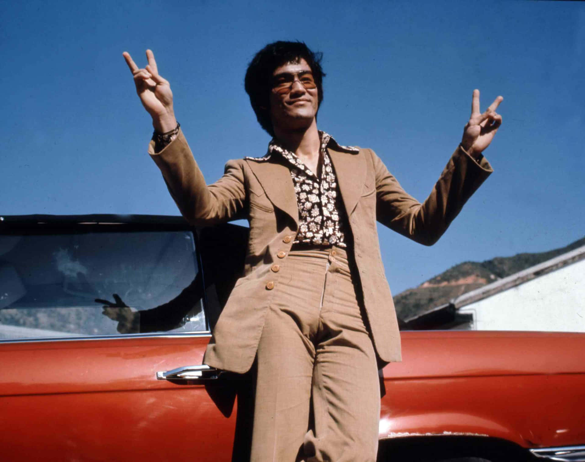 1970年代身穿棕色西裝的李小龍在紅色敞篷車旁比出和平手勢。 — 李小龍 壁紙