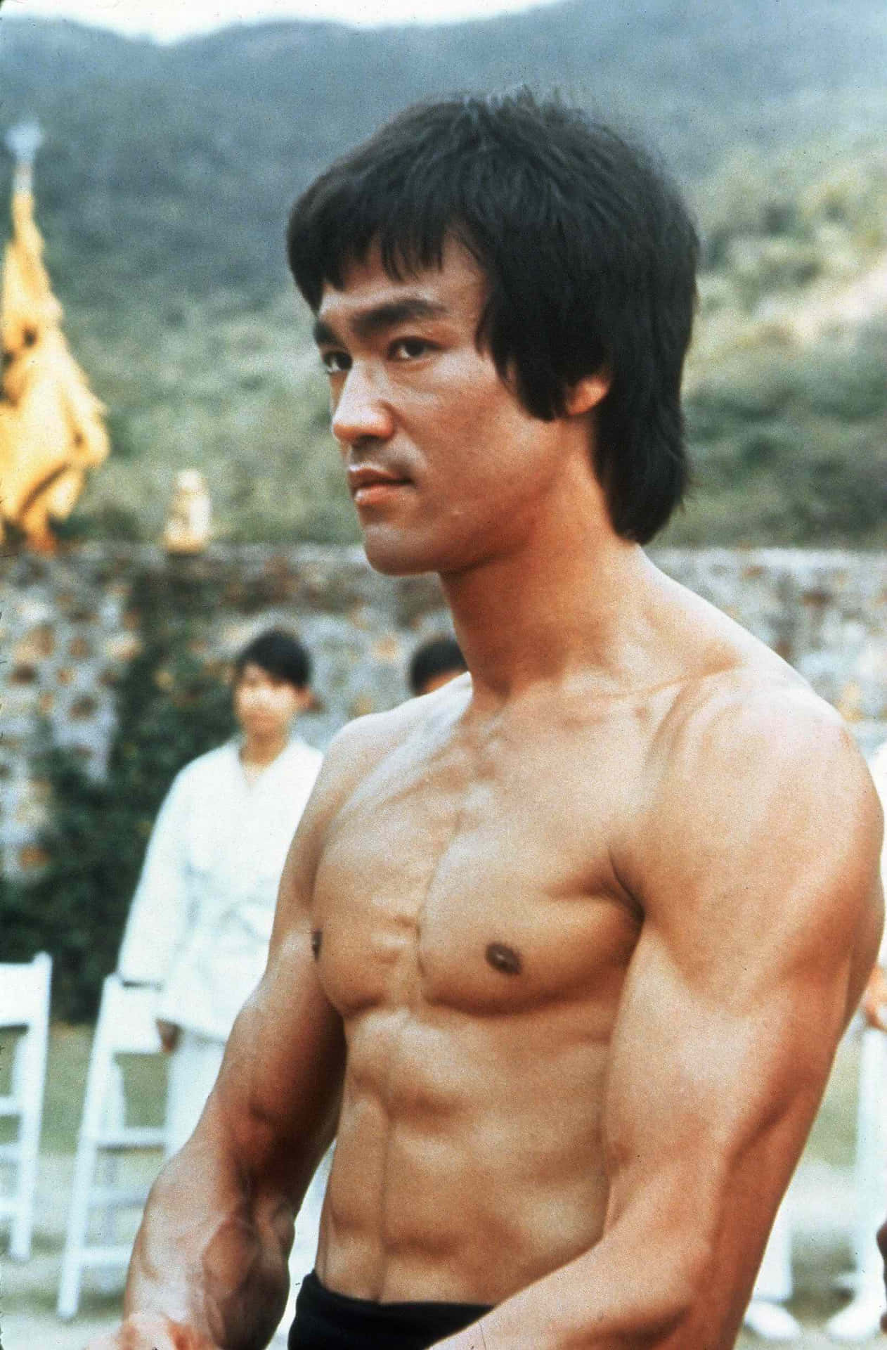 李小龍赤裸上身，肌肉發達，站在戶外靠近山脈的地方。 — 李小龍 壁紙