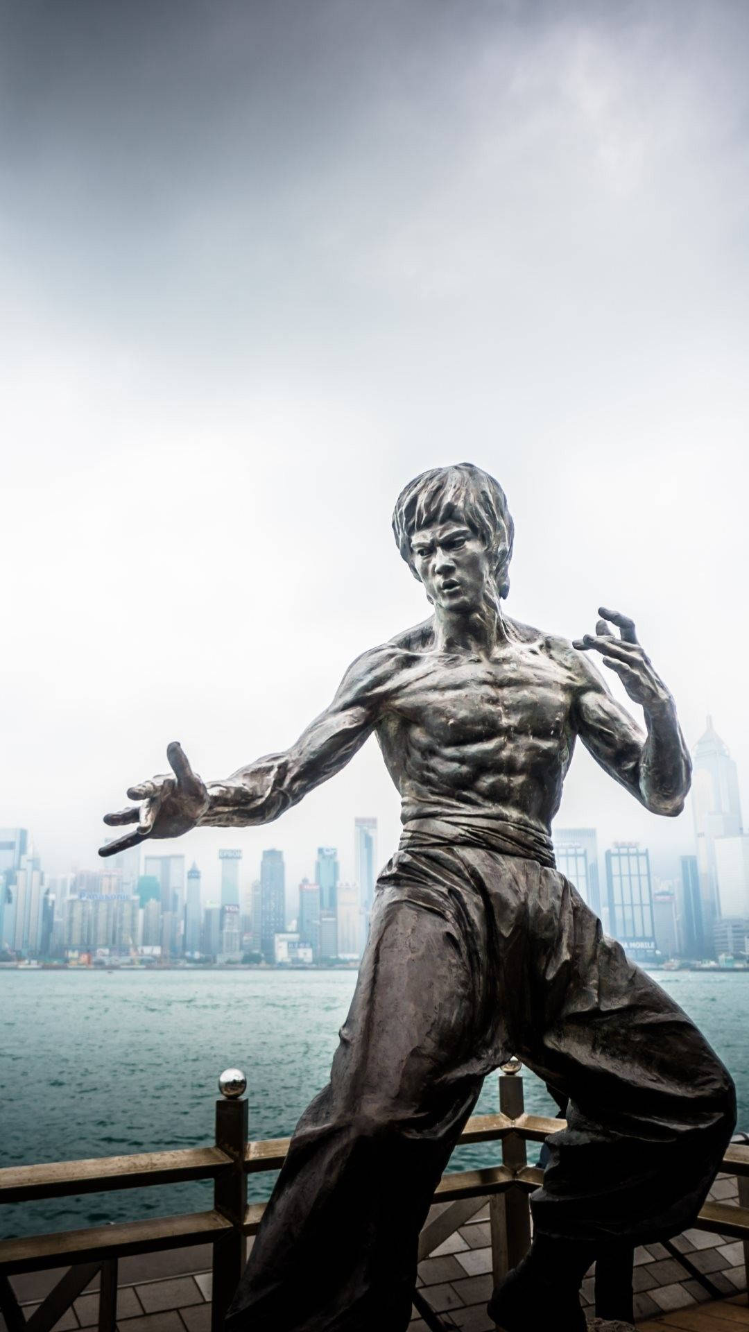 李小龍武術姿勢的青銅雕像，位於碼頭，背景為城市天際線。
