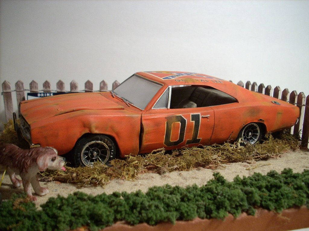 《杜克兄弟》經典賽車飛躍 壁紙 — 一架風化嘅橙色 Dodge Charger 模型，側面有「01」字樣，放喺乾草上面，旁邊有個木籬笆同一個細狗公仔。