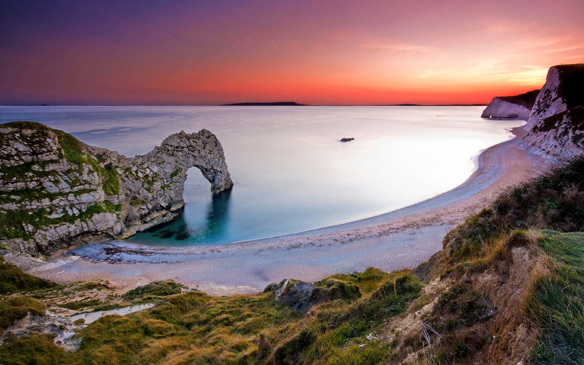 England 桌布 壁紙 — 杜德爾門天然石灰岩拱門在平靜的碧綠海洋和沙灘上，在熾熱的日落天空下閃耀。