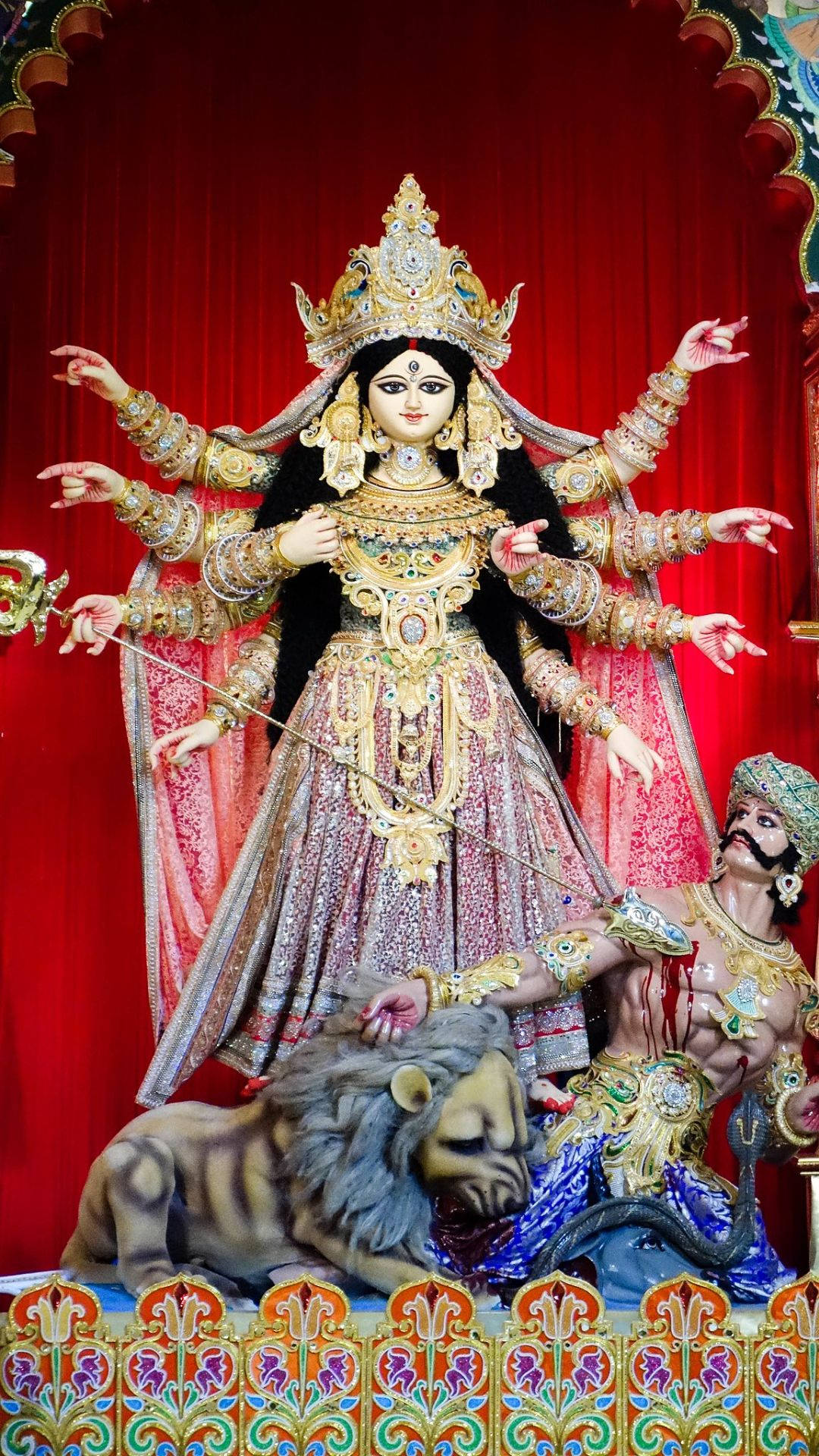 杜爾加女神 HD 與獅子相伴 壁紙 — 一座威嚴的十臂金色女神像，在華麗的紅色帷幕背景下，勝利地立於被擊敗的惡魔和獅子之上。