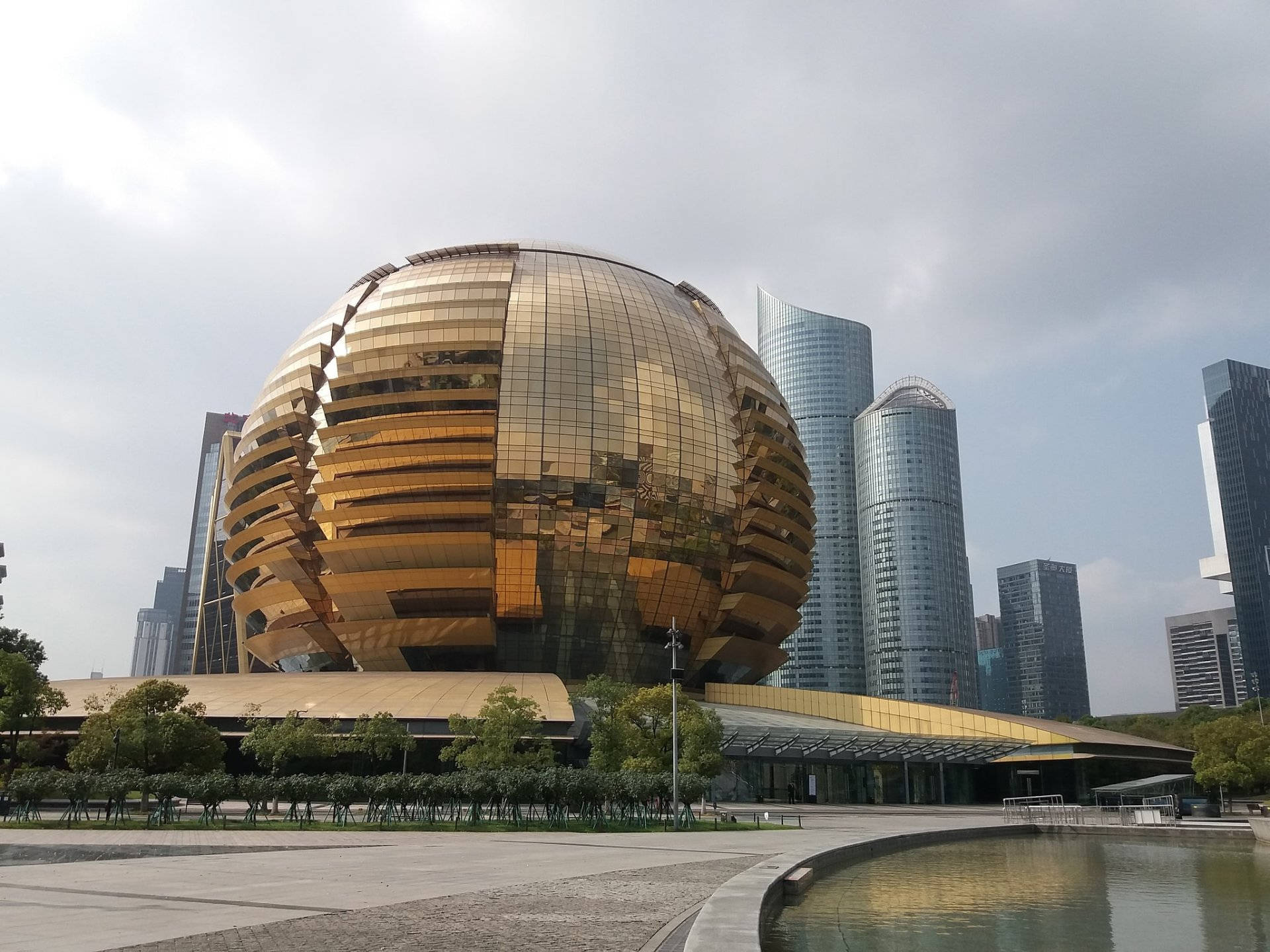 杭州國際會議中心 壁紙 — 一座巨大的金色球形建築，帶有反光面板，在陰天天空下，矗立在現代城市景觀中，周圍環繞著高聳的玻璃摩天大樓。