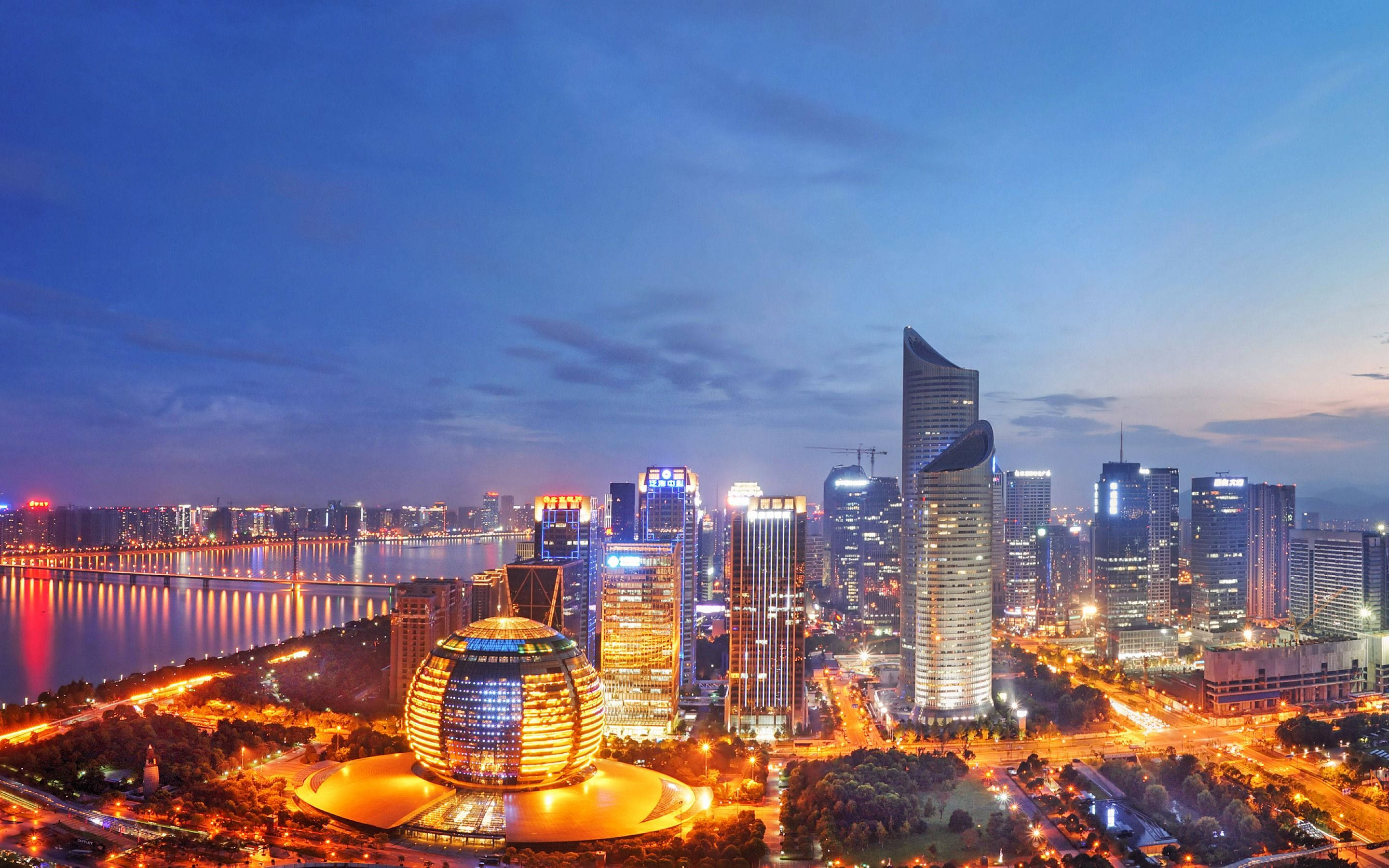 杭州城市夜景 壁紙 — 一個令人驚嘆的黃昏城市景觀，閃爍的摩天大樓和一條在深藍色天空下反射溫暖燈光的河流。