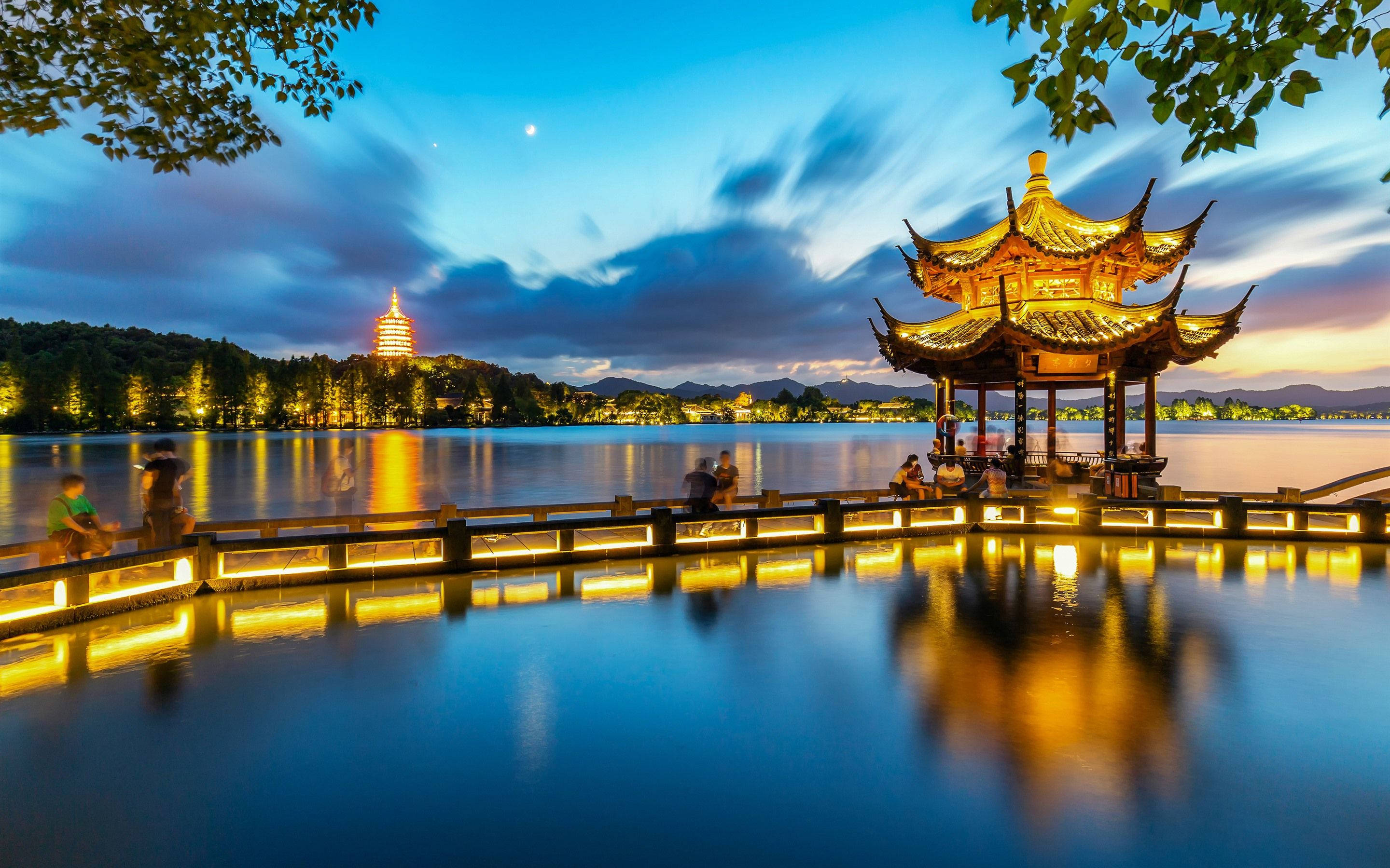 杭州湖景 壁紙 — 月夜下，平靜湖面倒映著發光的傳統亭閣和遠處寶塔，一片寧靜的黃昏湖邊景色。