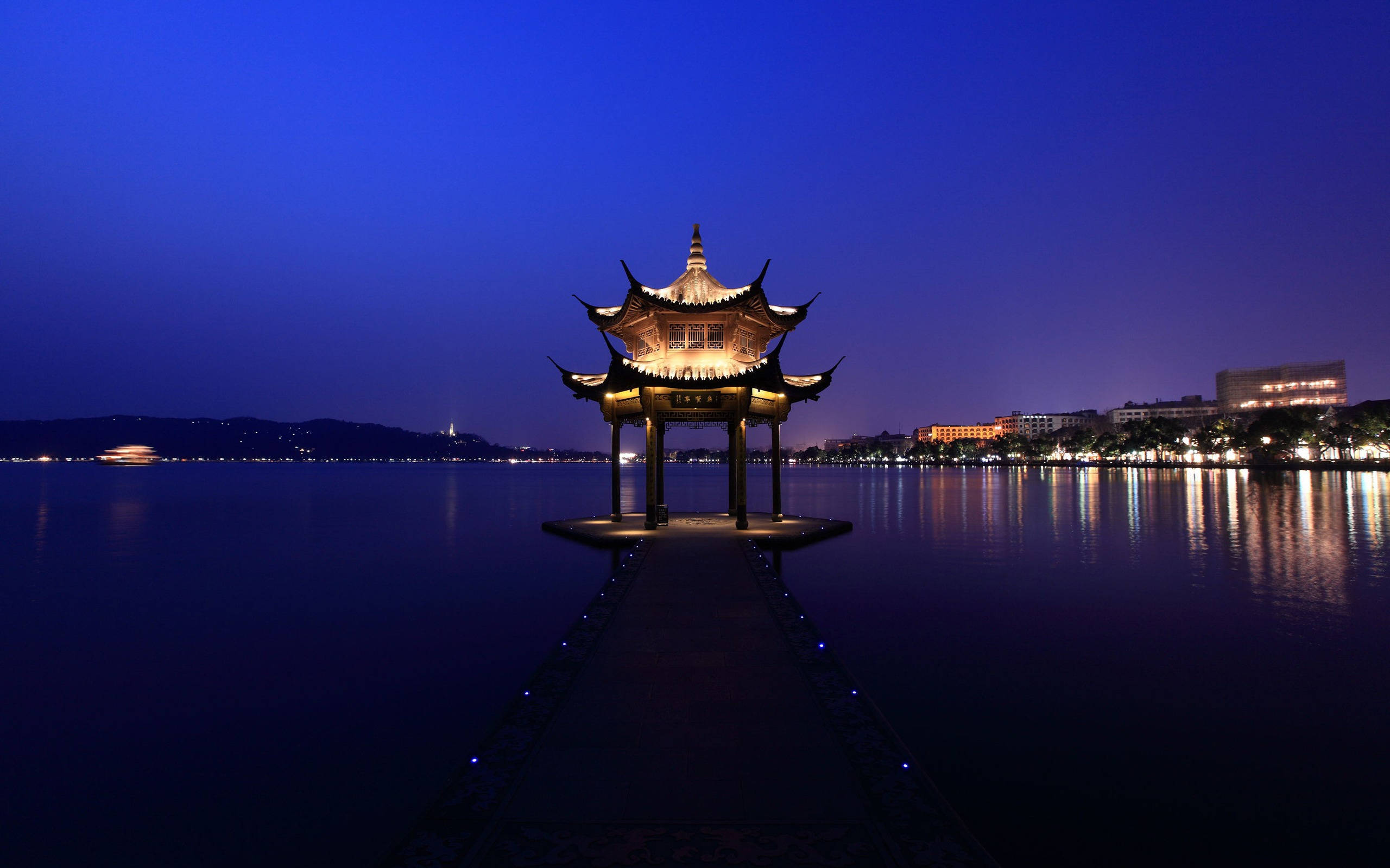 杭州西湖 壁紙 — 一座發光的傳統中式亭子矗立在寧靜湖面上的碼頭盡頭，在深藍色的暮色天空下。