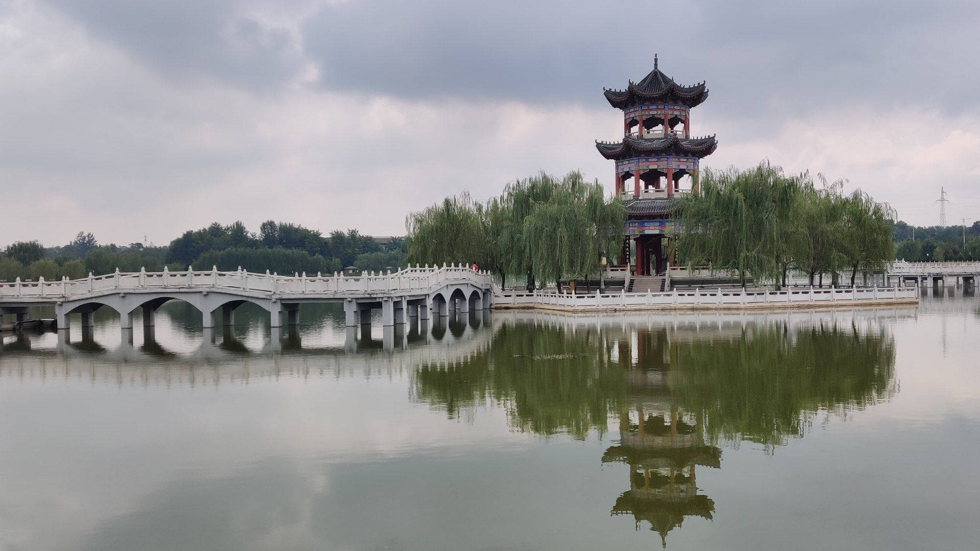 杭州西湖日落美景 壁紙 — 一座傳統中式亭閣和石橋在陰天之下平靜地倒映在寧靜的水面上。