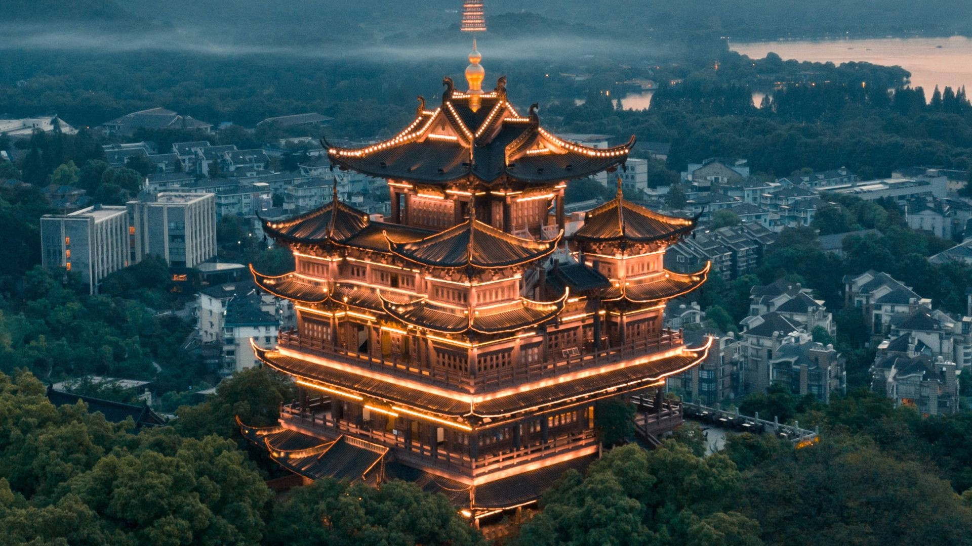 杭州西湖日落風景 壁紙 — 一座雄偉的傳統中式寶塔在燈光下溫暖地發光，映襯著霧氣瀰漫的暮色天空，下方是密集的城市景觀。