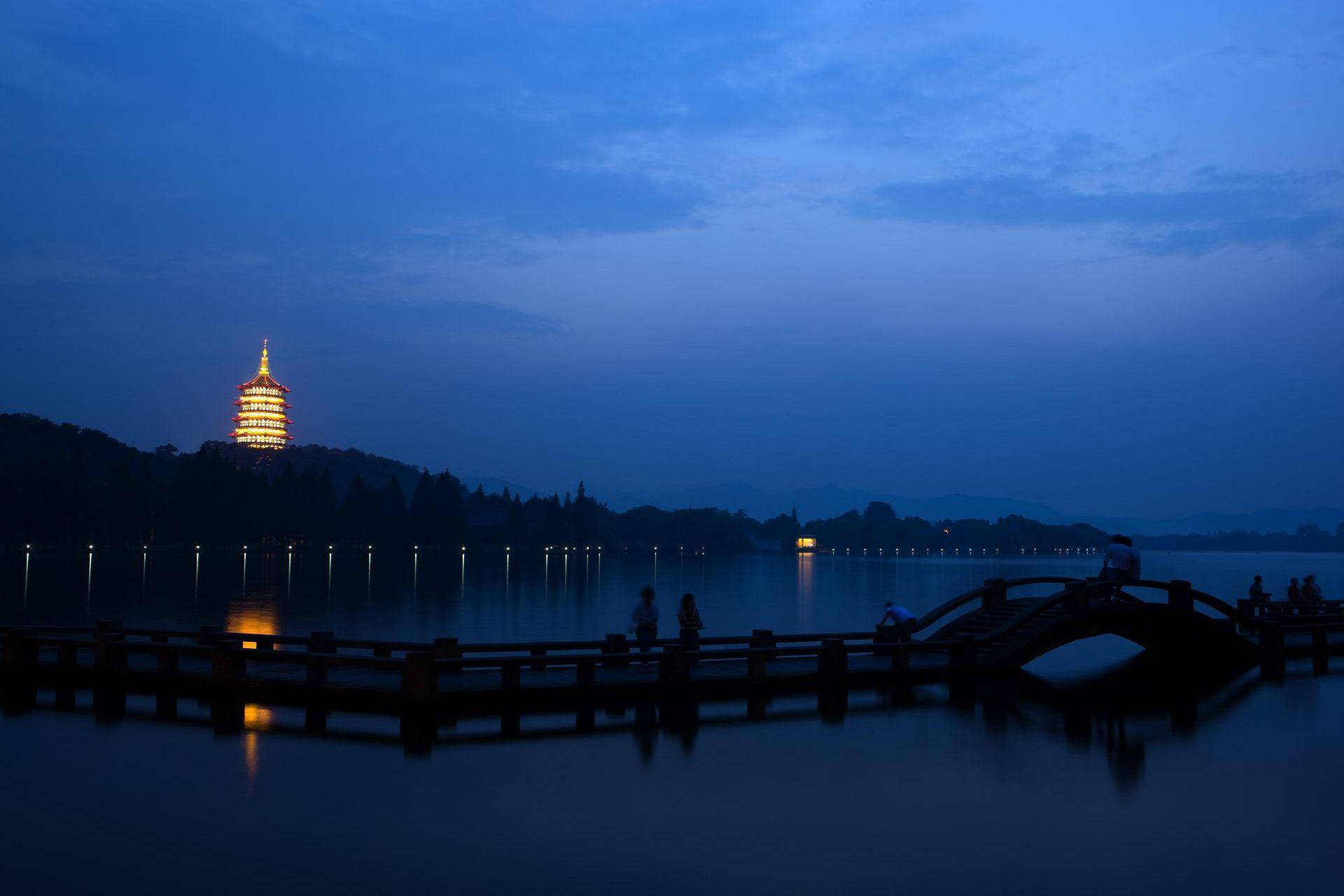 杭州西湖晨曦 壁紙 — 寧靜的黃昏場景，山丘上發光的寶塔倒映在平靜的水面上，木橋上的人影剪影。