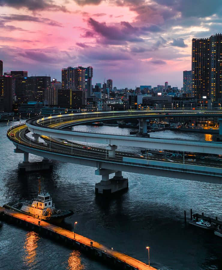 東京odaiba的活力彩虹橋 壁紙 — 一條蜿蜒的高速公路橋在黃昏時分彎曲在黑暗的水面上，粉紫色雲彩和城市燈光倒映在水面。