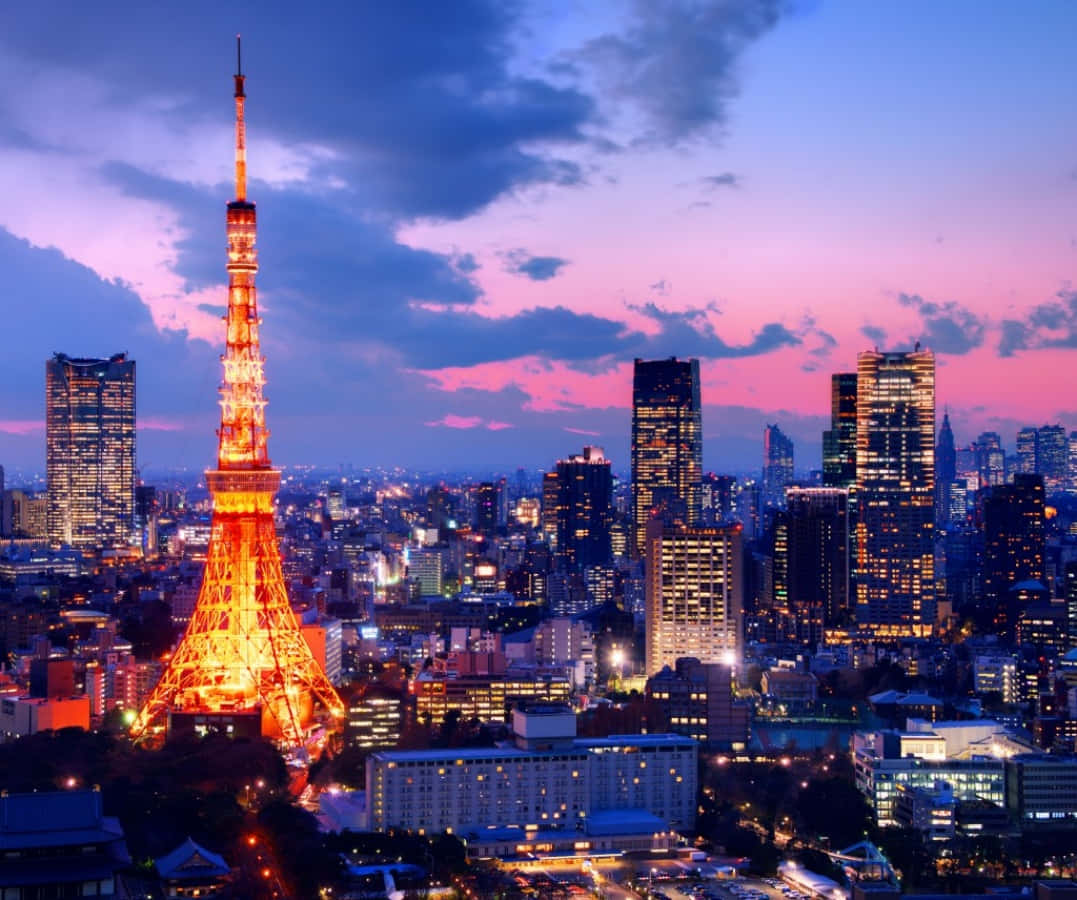 東京 桌布 壁紙 — 東京塔在暮色天空中溫暖地發光，天空佈滿粉紅和紫色的雲彩，下方是燈火通明的摩天大樓天際線。