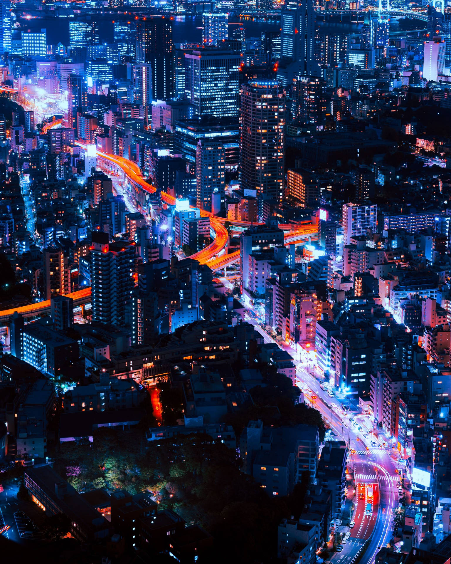 東京夜景全景，霓虹燈光交織 壁紙 — 夜晚霓虹燈照亮城市的鳥瞰圖，有發光的藍色摩天大樓和橙色的車流燈光軌跡。