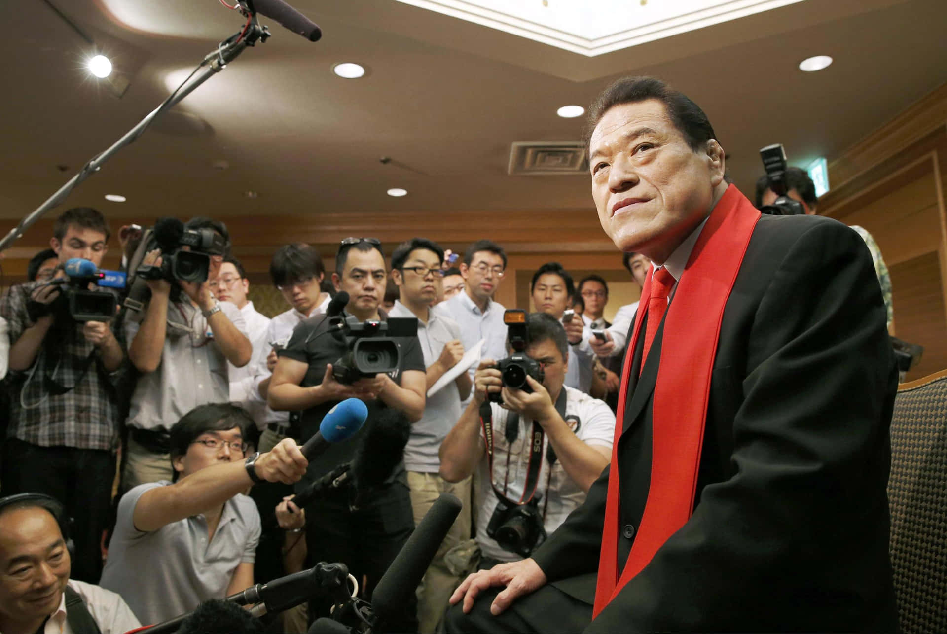 室內，身穿黑色西裝和紅色圍巾的男子被記者和攝影機包圍。