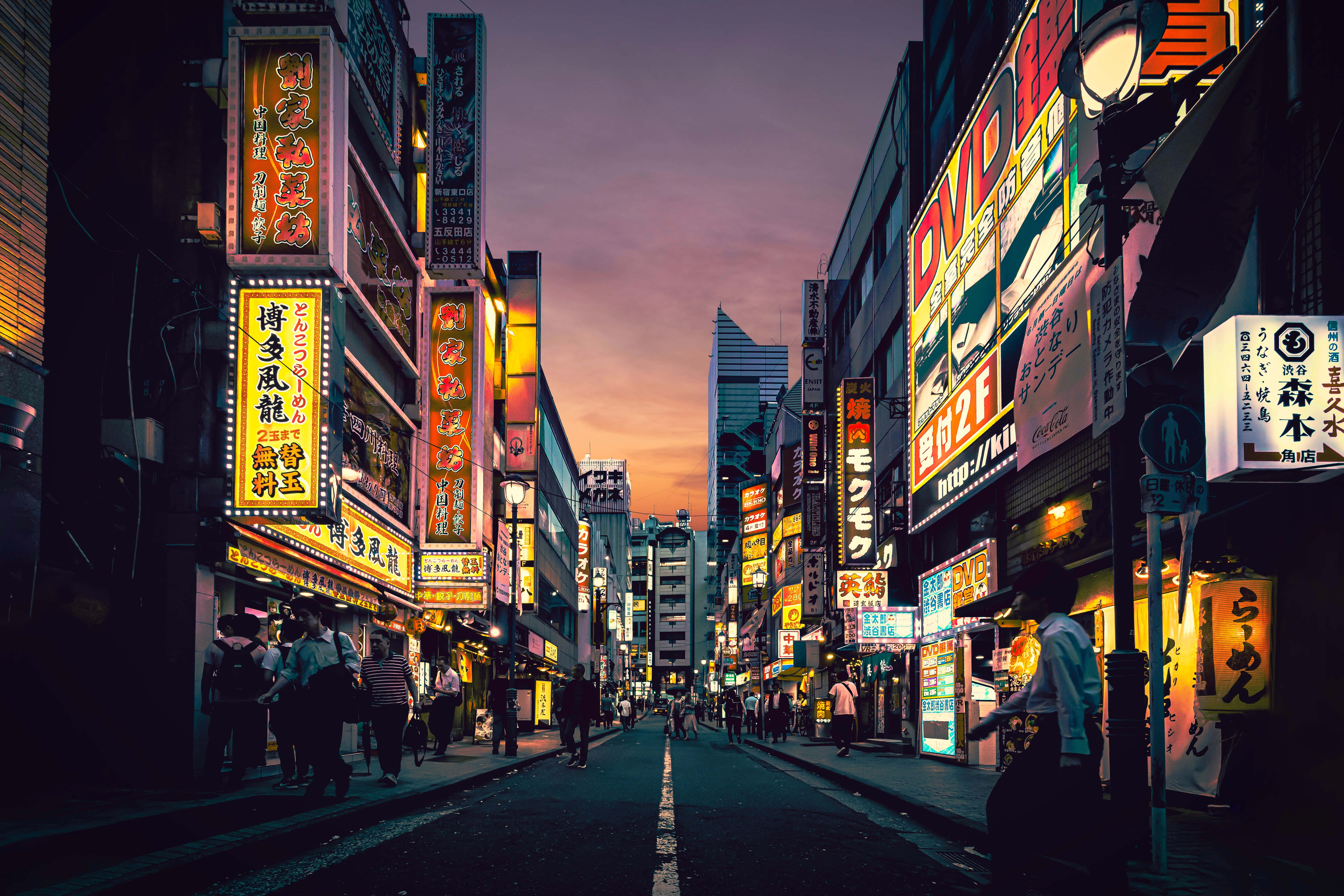東京日本霓虹燈的活力場景 壁紙 — 黃昏時分，東京一條繁忙的小巷，發光的霓虹燈招牌映照在濕漉漉的人行道上，行人走在高聳的建築物下。