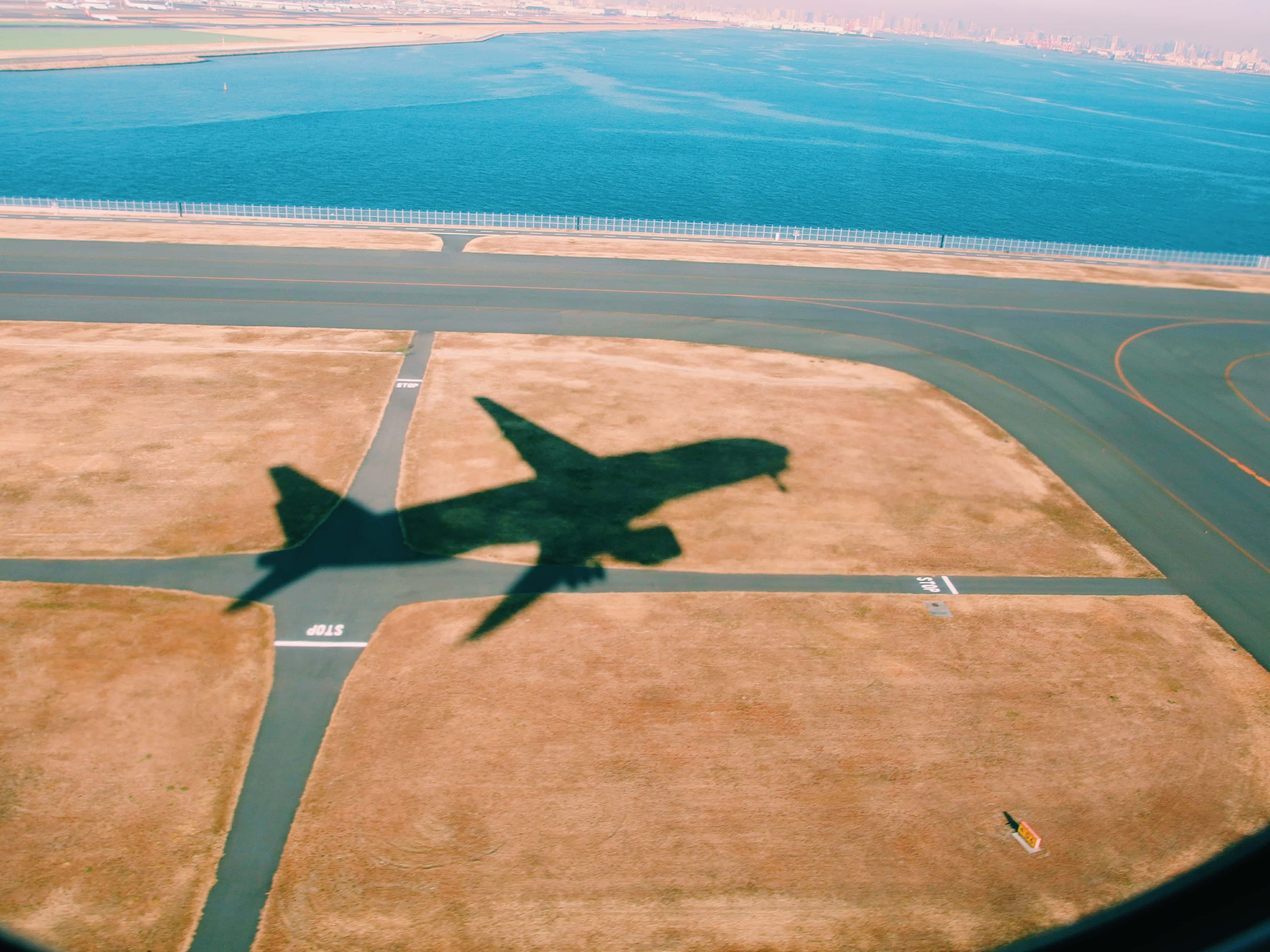 東京羽田機場飛機起飛時的影子 壁紙 — 從空中俯瞰大型飛機影子投射在跑道和乾草上，旁邊是碧綠色的海洋和遠處的城市天際線。
