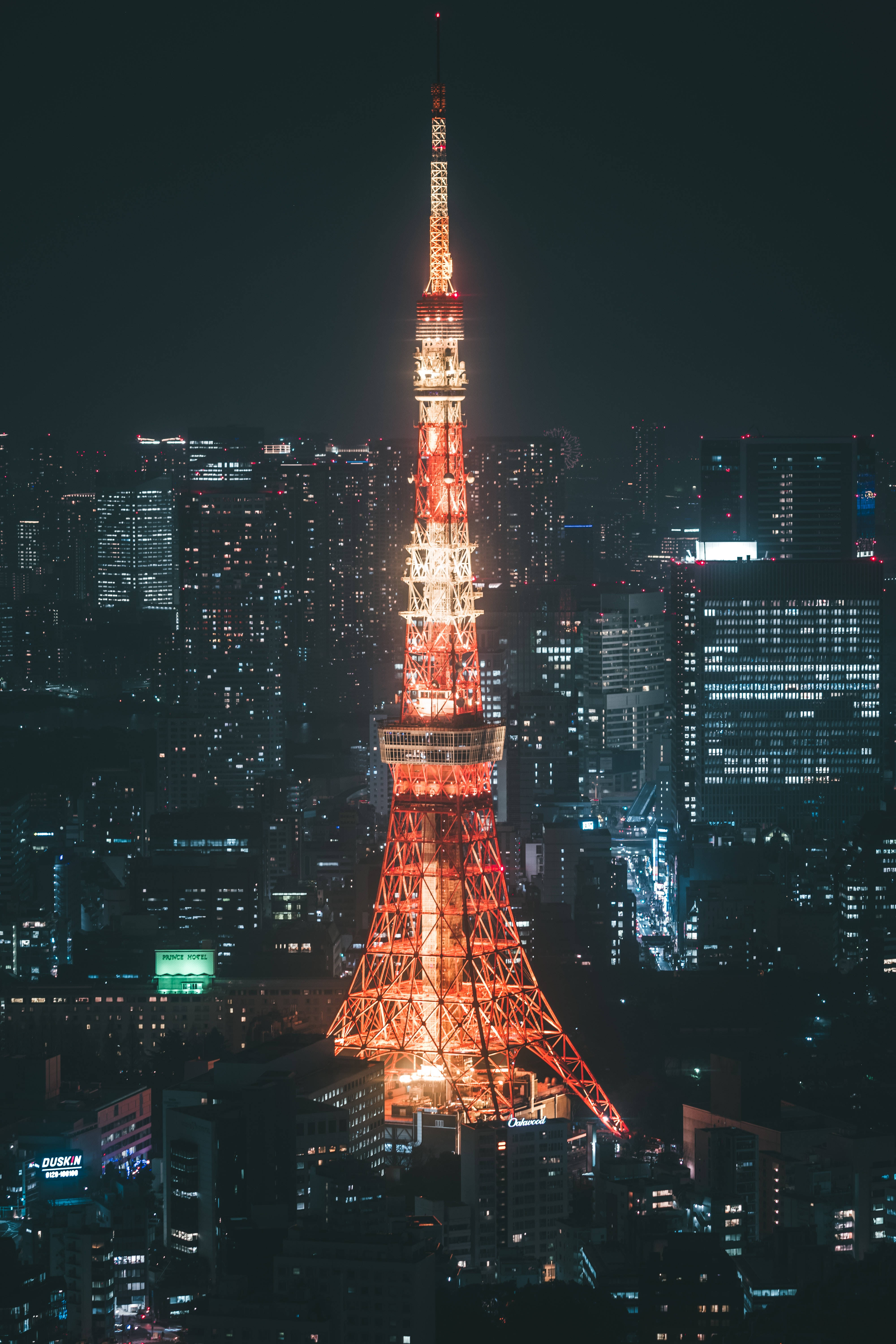 東京鐵塔紅白燈光夜景 壁紙 — 東京塔在黑暗的天空中溫暖地發光，周圍是夜晚燈火輝煌的摩天大樓閃爍的城市景觀。