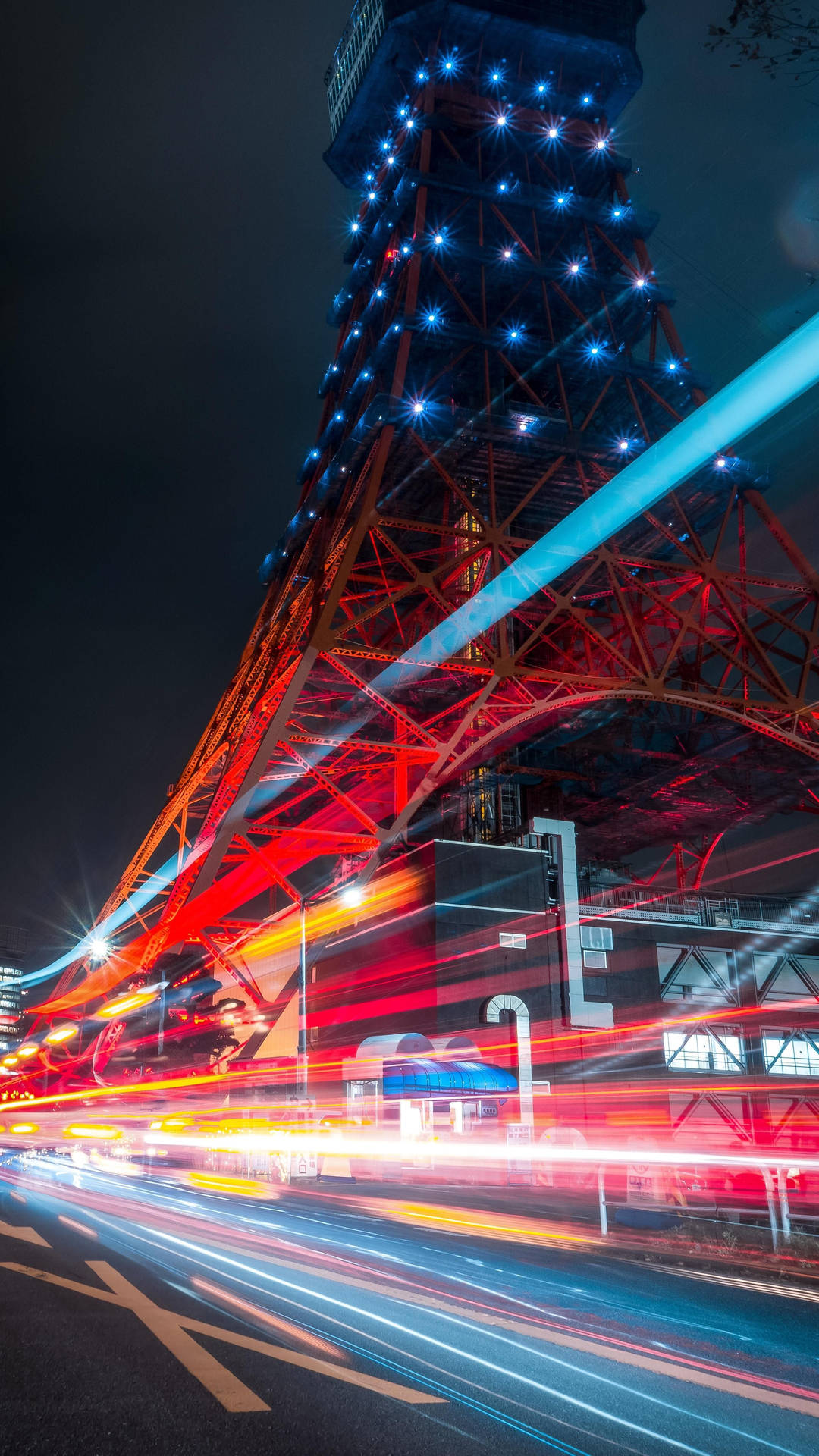 東京鐵塔車燈拖影 壁紙 — 東京鐵塔在夜晚閃耀紅藍光芒，下方車流形成光軌的充滿活力的夜景照片。
