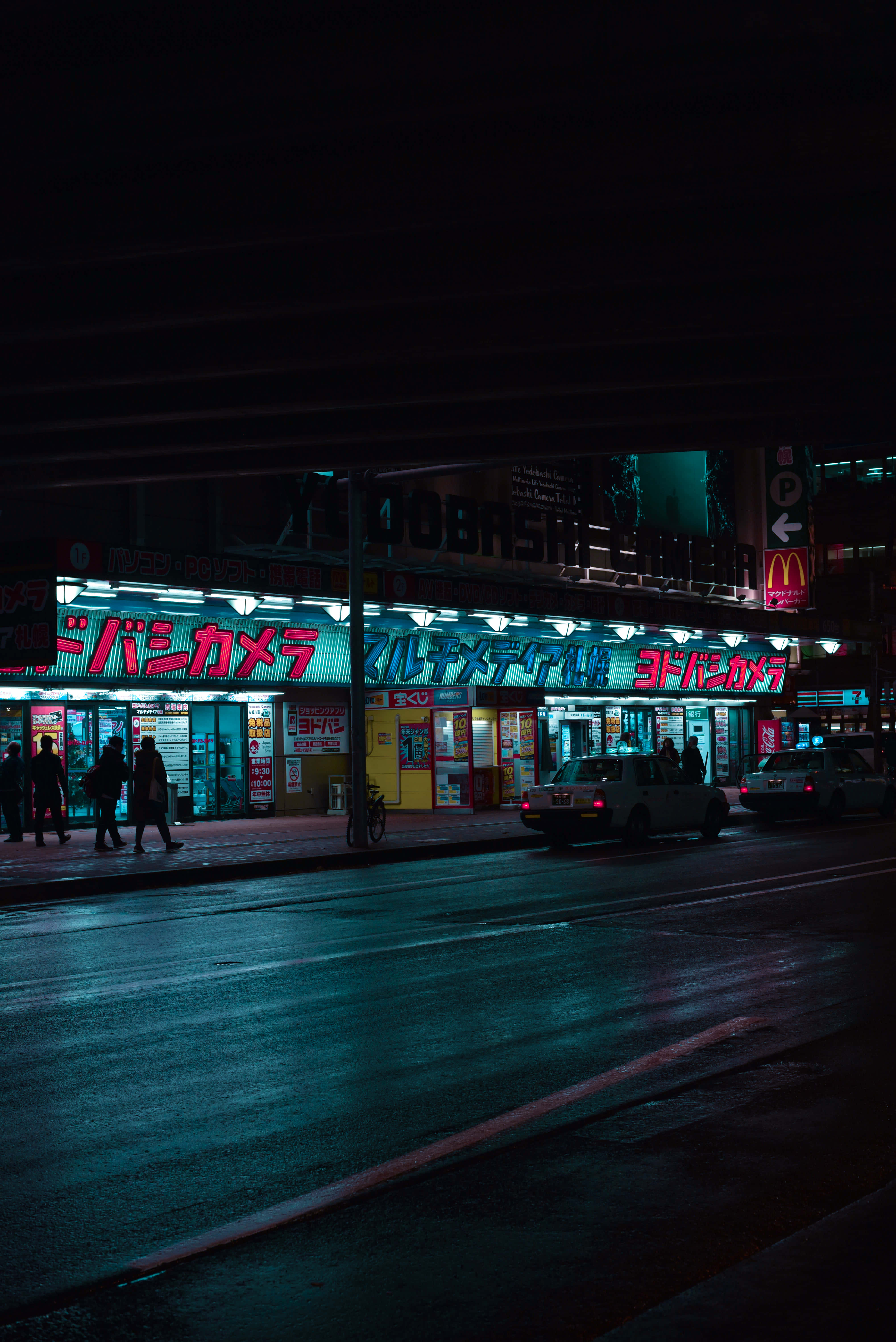 Vibes 桌布 壁紙 — 東京雨夜的街道，被閃爍的霓虹燈照亮，倒映在濕漉漉的路面上，有行人和的士經過。