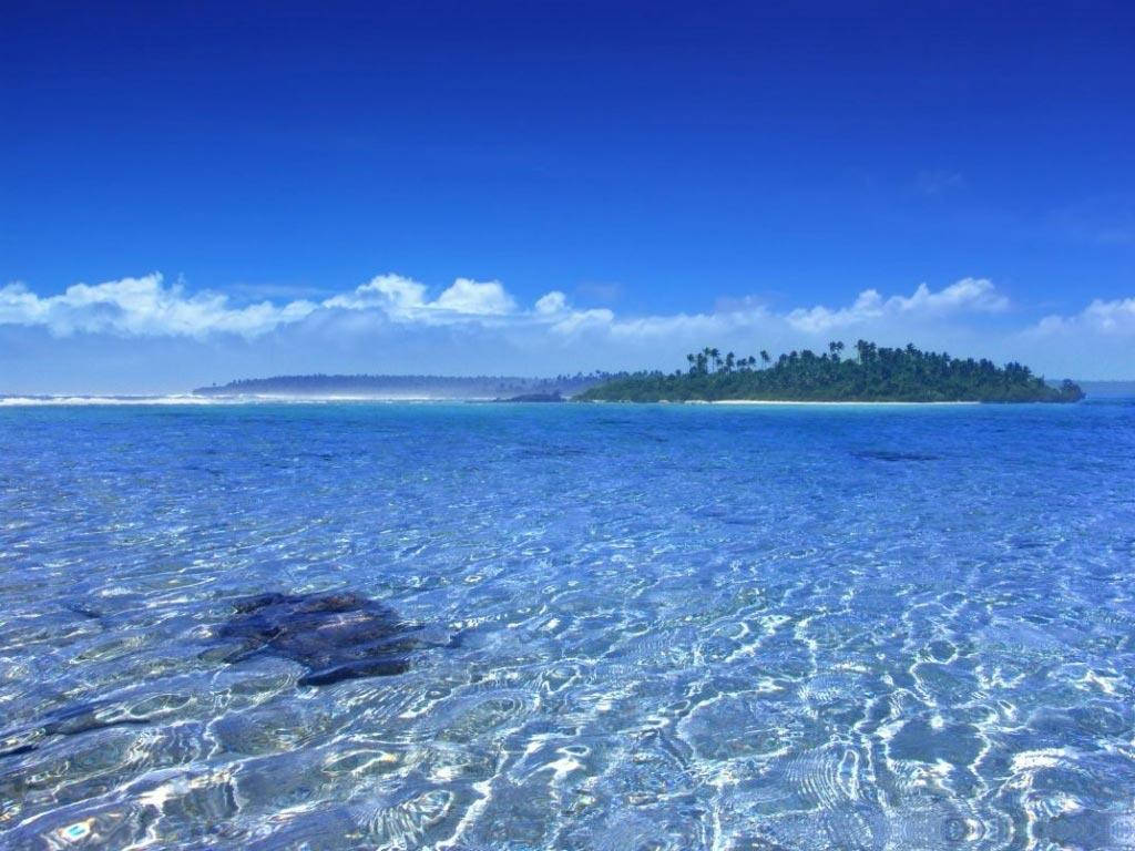 東加淺藍清澈海域 壁紙 — 清澈的碧綠海水泛起漣漪，流向一個鬱鬱蔥蔥的綠色島嶼，天空湛藍，白雲點綴。