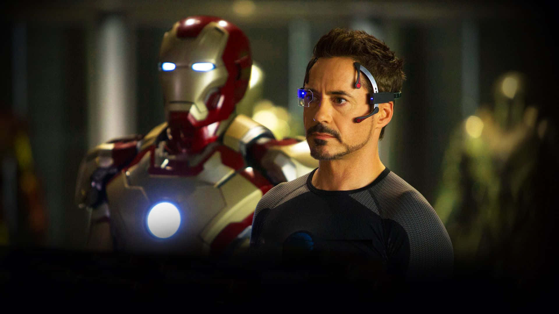 Iron Man Movies 桌布 壁紙 — 東尼·史塔克在他的實驗室中，身後有發光的鐵甲奇俠戰衣，處於昏暗的未來感燈光下。
