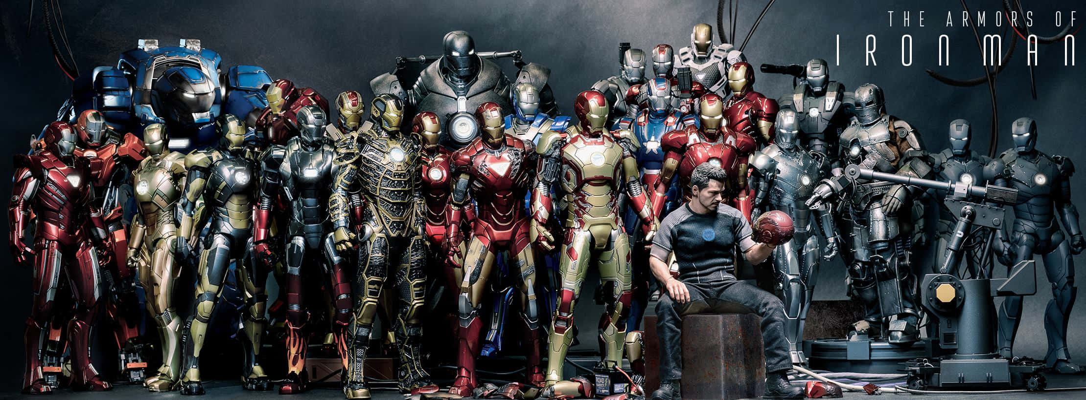 Iron Man Suit 桌布 壁紙 — 東尼·史塔克坐在燈光昏暗的工作室中，周圍環繞著大量細節精緻的鐵甲奇俠戰甲。