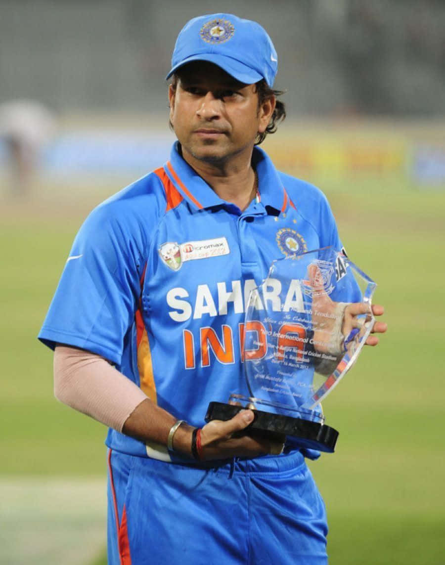 Sachin Tendulkar 桌布 壁紙 — 板球傳奇沙欽·坦杜卡身穿藍色印度球衣，在球場燈光下的綠色場地上手持玻璃獎盃。