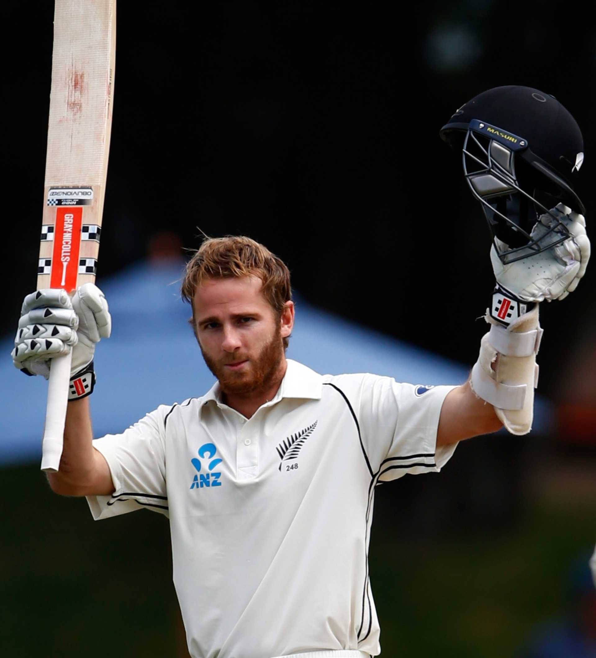 板球明星 Kane Williamson 壁紙 — 一名身穿白色球衣的板球運動員在模糊的體育場背景下，高舉球棒和頭盔慶祝勝利。