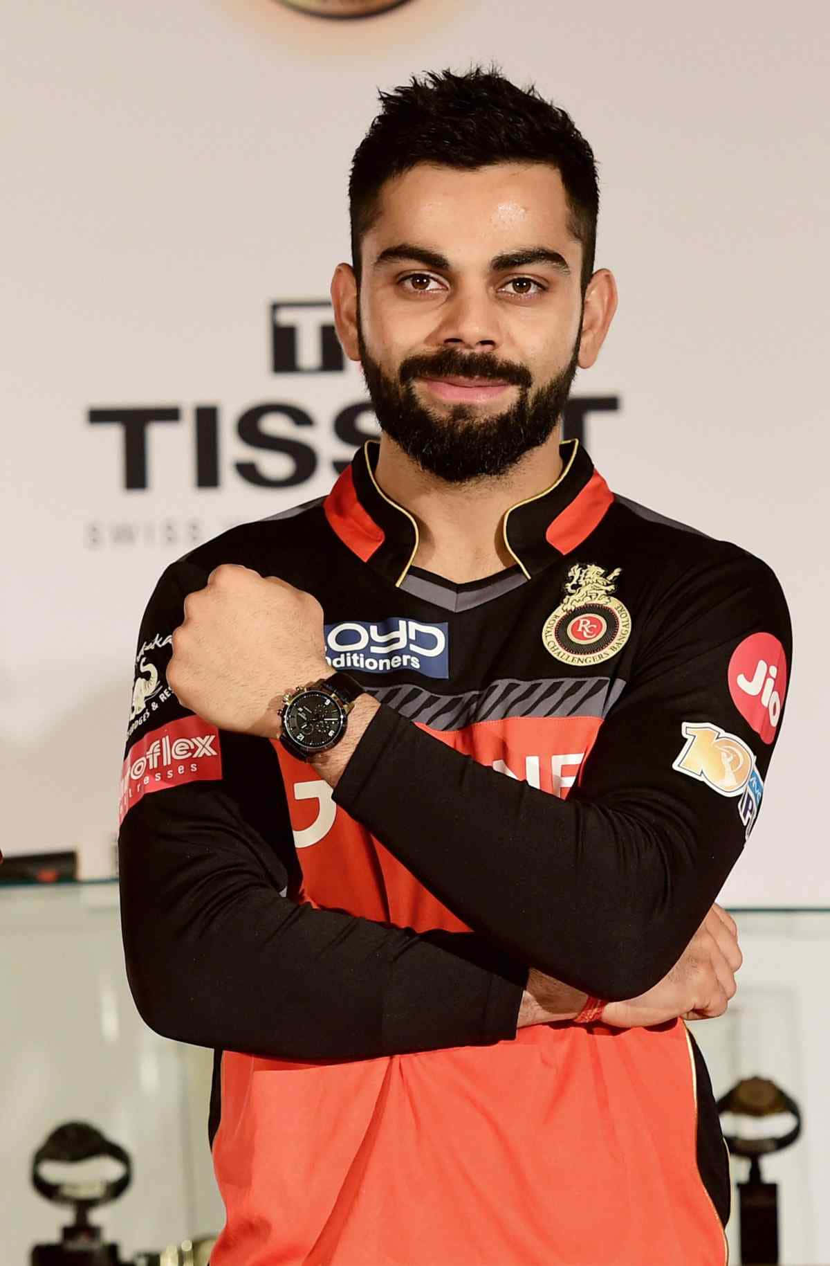 Rcb 桌布 壁紙 — 板球明星 Virat Kohli 身穿黑色和橙色球衣，雙臂交叉，在品牌背景前自信地擺姿勢。