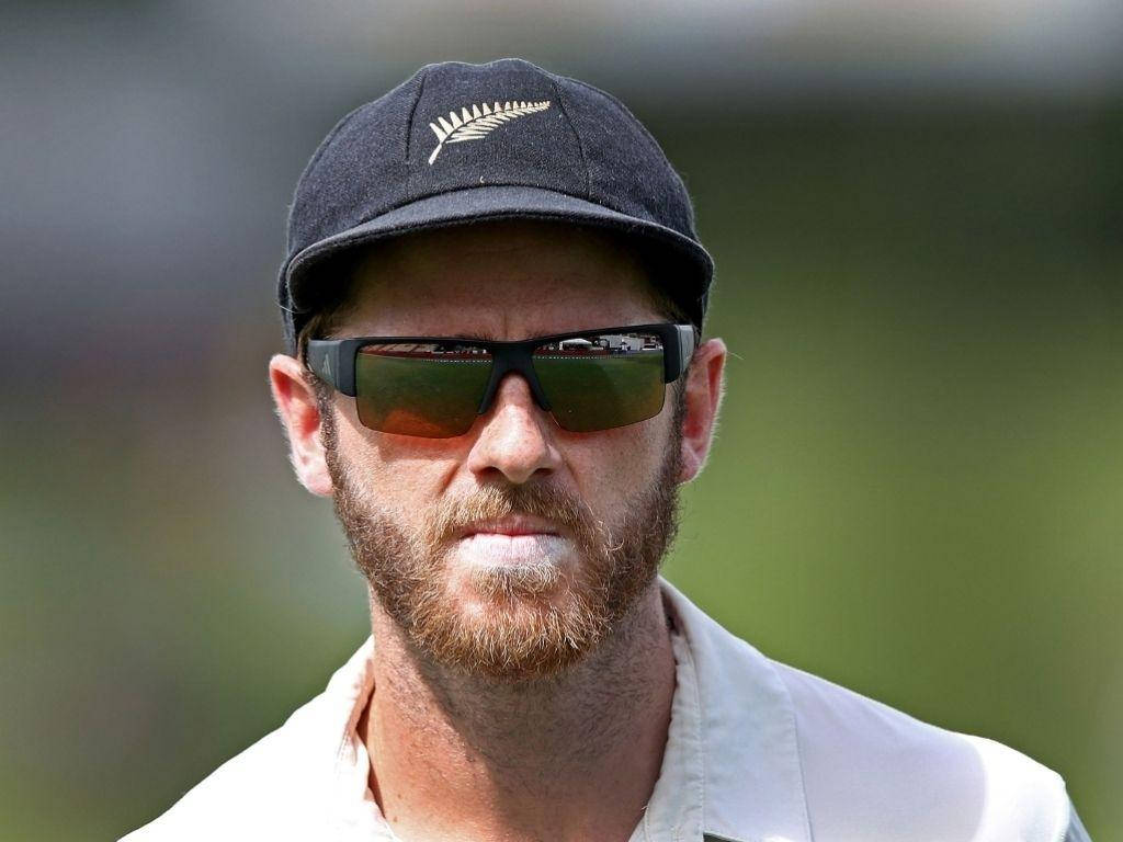 板球運動員 Kane Williamson 壁紙 — 一位專注的新西蘭板球運動員，留著鬍鬚並戴著太陽眼鏡，在模糊的綠色場地上凝視。