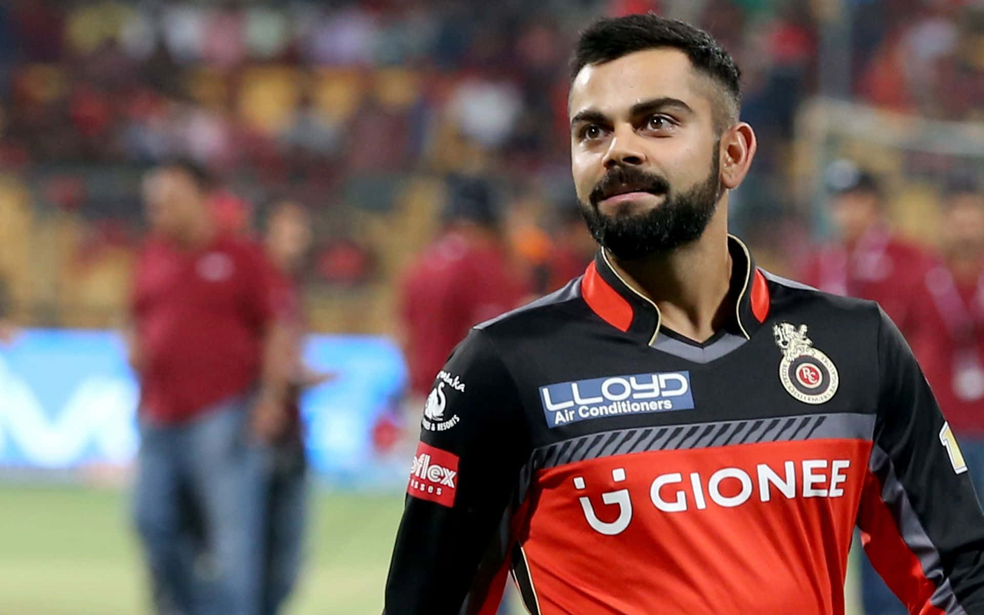 Rcb 桌布 壁紙 — 板球運動員 Virat Kohli 身穿紅黑球衣，自信地站在燈光明亮的體育場上，背景是模糊的觀眾。