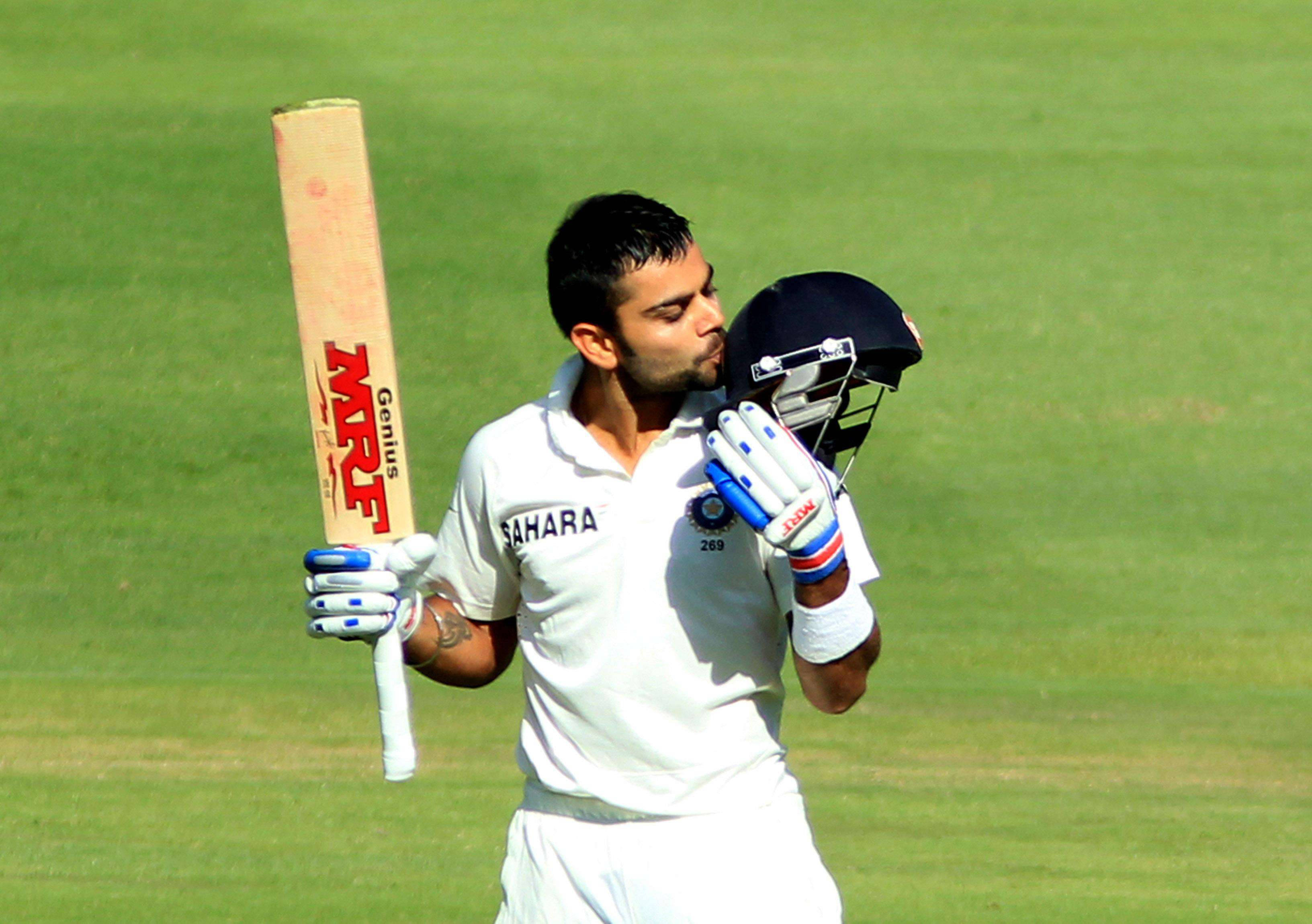 皇家挑戰者班加羅爾 桌布 壁紙 — 板球運動員 Virat Kohli 身穿白色制服，在陽光明媚的綠色場地上手持球棒和頭盔。