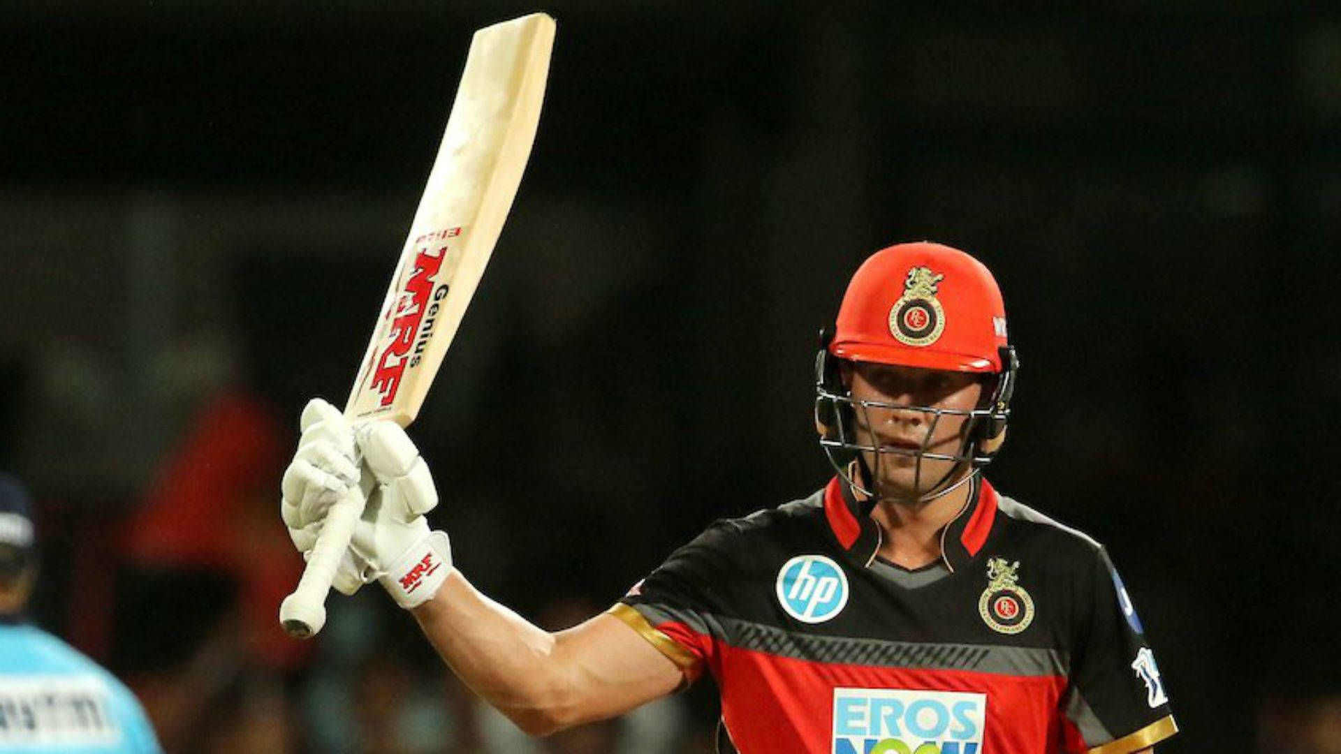 royal challengers bangalore 桌布 壁紙 — 板球運動員戴著紅色頭盔和黑色球衣，在體育場燈光下勝利地舉起木製球棒。