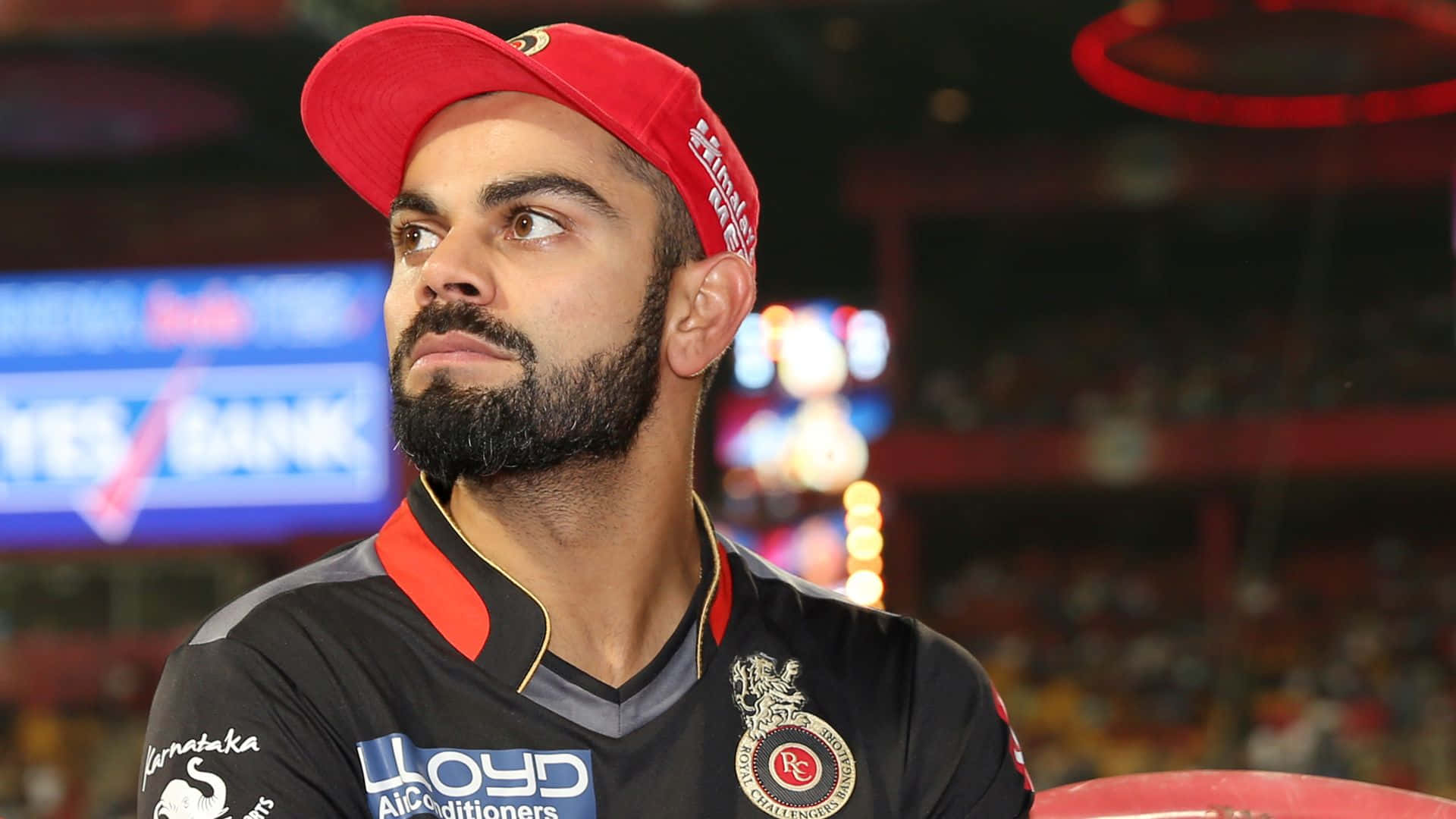 Virat Kohli 桌布 壁紙 — 板球運動員維拉·哥利戴上紅色帽子和黑色球衣，若有所思地凝視著身後的球場燈光。