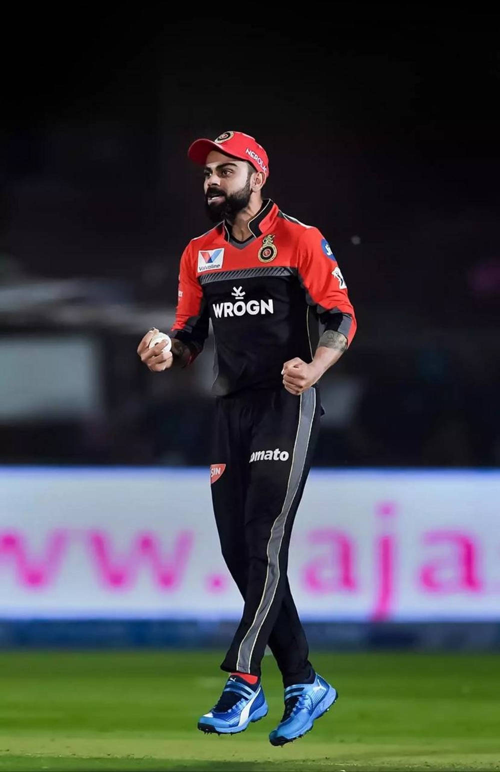 virat kohli rcb 桌布 壁紙 — 板球運動員維拉特·哥利戴著紅色帽子和穿著紅色球衣，在體育場燈光下的綠色場地上邁步。