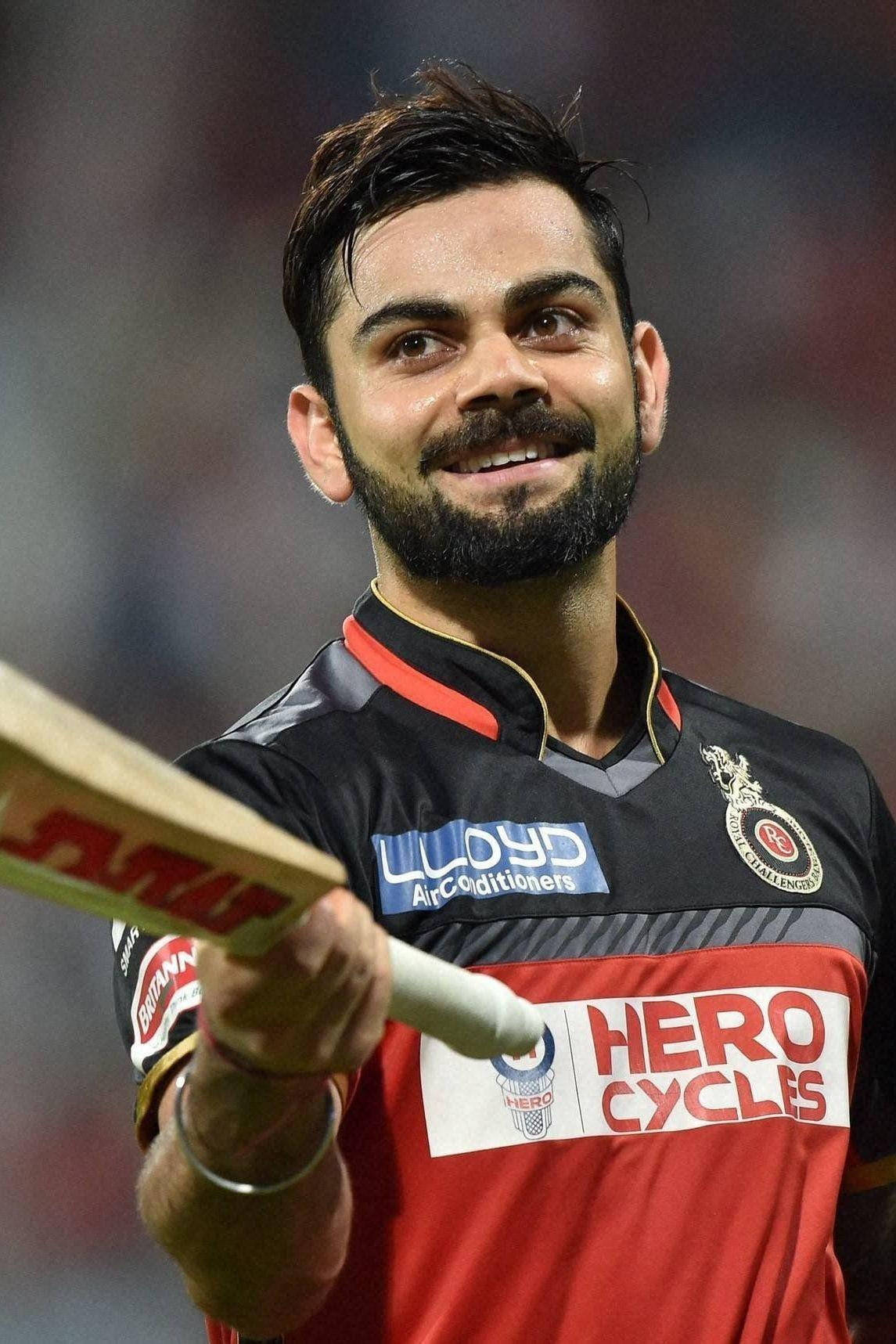 virat kohli rcb 桌布 壁紙 — 板球運動員維拉特·科利在明亮燈光下的體育場內自信地微笑，手持球棒。