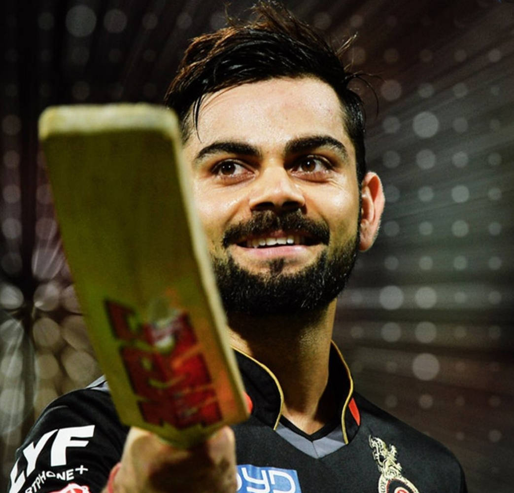 royal challengers bangalore 桌布 壁紙 — 板球運動員維拉特·科利在模糊的體育場背景下自豪地微笑著拿著球棒。