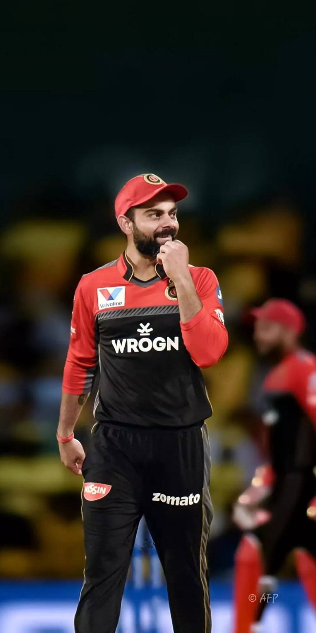 virat kohli rcb 桌布 壁紙 — 板球隊隊長維拉·哥利戴上紅色帽子和黑色球衣，在夜間比賽的球場燈光下，手托下巴沉思。