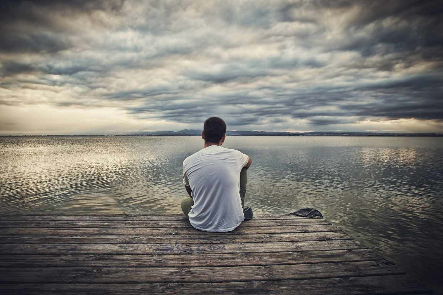 一個男人獨自坐在木碼頭上，俯瞰著多雲天空下的平靜湖泊。