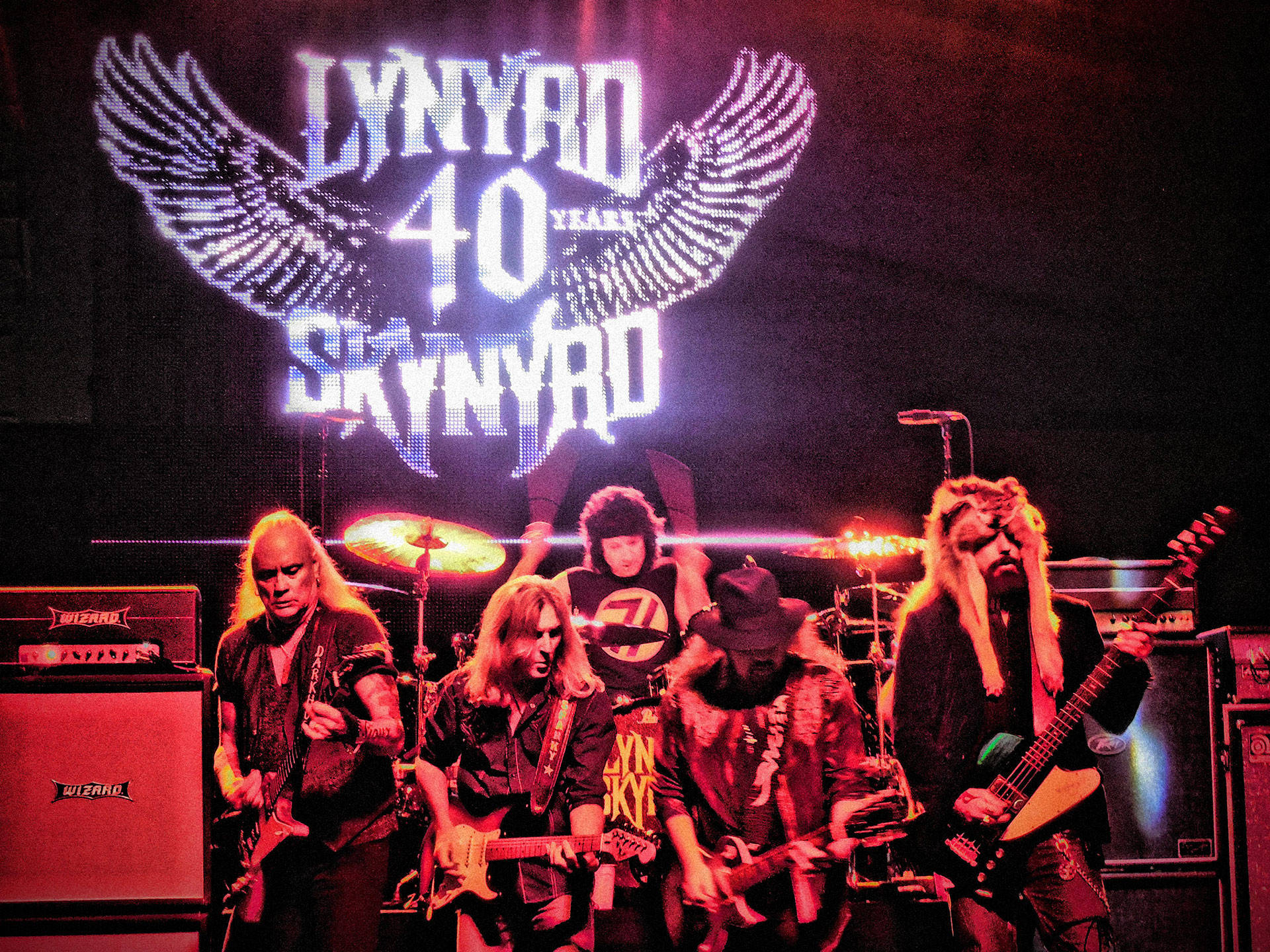 Lynyrd Skynyrd 樂隊在舞台上現場表演，身後有發光的翅膀標誌。
