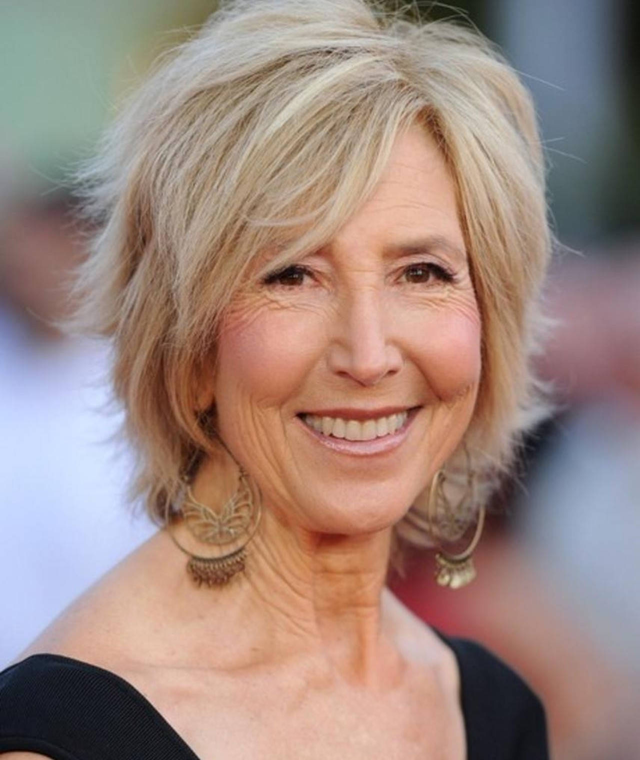一位微笑的金髮女性戴著圈耳環，身穿黑色上衣。