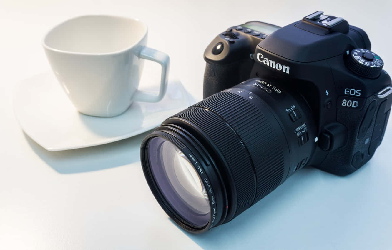 黑色 DSLR 相機旁邊是白色咖啡杯放在碟上，置於白色表面。 — 相機 壁紙